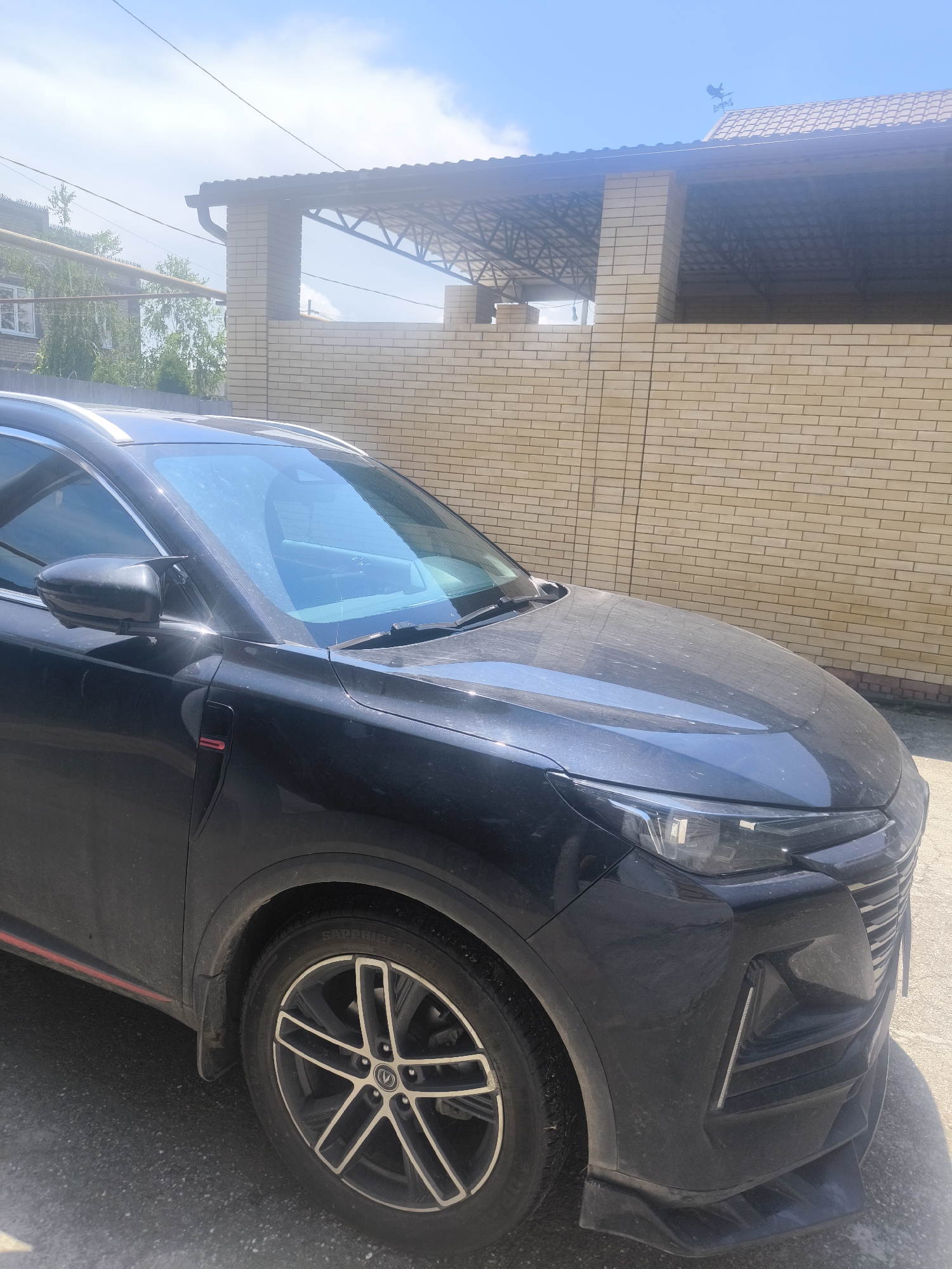 The image displays the front-left section of a dark-colored SUV, likely black or dark grey, parked outdoors in the city of Unknown, Unknown. The vehicle is coated with a visible layer of dust or dirt across its surfaces, including the hood, windshield, and side panels.
The SUV features modern headlights with an integrated horizontal LED strip and a prominent front grille characterized by vertical chrome-like slats. The lower bumper includes a dark-colored spoiler-like extension and vertical daytime running lights or vents on either side. A red accent strip is visible along the lower part of the front quarter panel, extending towards the wheel well. The front wheel is equipped with a five-spoke alloy rim, dark-colored with polished silver accents on the spokes, and the tire sidewall displays "SAPPHIRE SVR H/T" in white lettering. A black side mirror is visible, partially reflecting bright sunlight. Silver-colored roof rails run along the top edges of the vehicle. A red badge or accent is present on a black trim piece near the front fender/door seam.
In the background, to the immediate right of the vehicle, a light-colored (tan/beige) brick wall extends horizontally. Above this wall, a dark-colored, sloped roof structure with visible metal truss supports provides a covered area, possibly a carport or patio. A dark rain gutter is attached to the edge of this roof. Further back and to the right, a tiled roof with a bird-shaped weathervane is discernible on a distant building.
To the left of the vehicle, in the mid-ground, another brick building is visible, featuring a window with a white frame. Green foliage from trees and bushes is present behind this building and in the center-left background. Utility lines traverse the upper-left portion of the image against a bright blue sky with scattered wispy white clouds, indicating a clear and sunny day. The ground beneath the vehicle appears to be an asphalt or paved surface, also showing signs of dust. No people are visible in the frame.