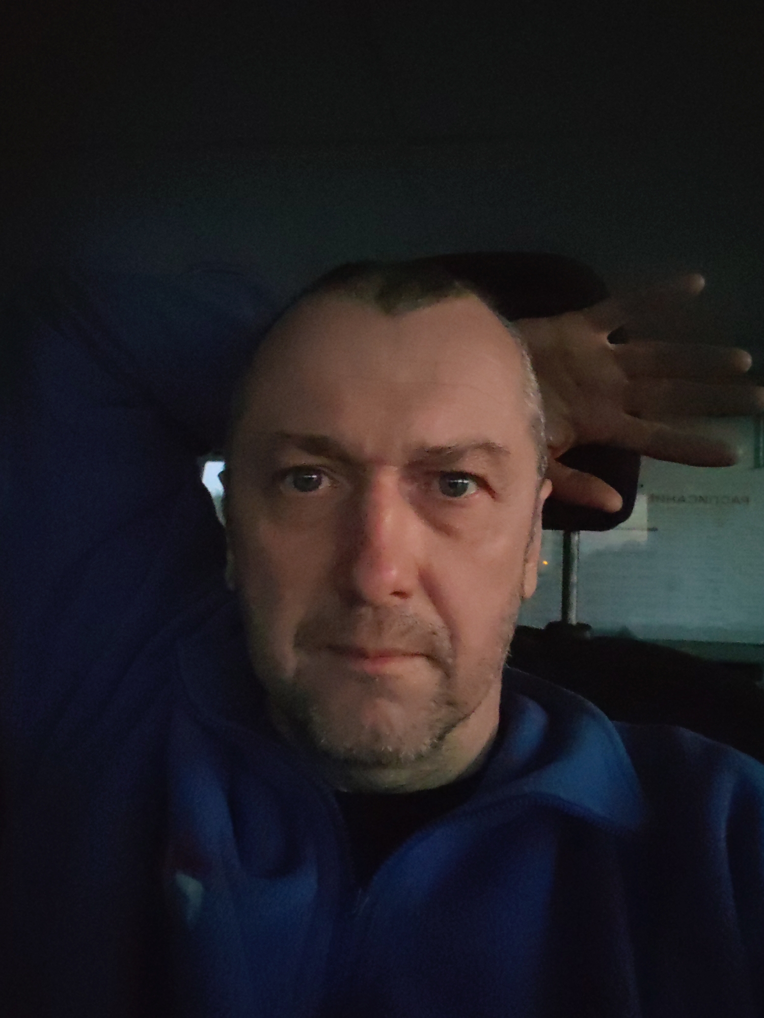A middle-aged man is taking a selfie in a dimly lit environment. He is wearing a blue jacket. His right arm is raised, with his hand near his head. There is an object that is possibly a screen or sign with lettering on it visible behind him. The location details, including the city and country, are "Unknown, Unknown."