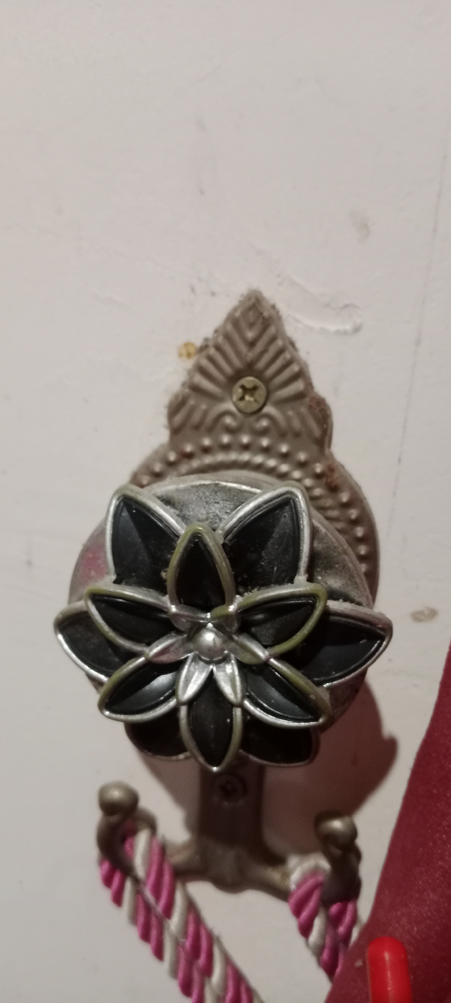 A decorative metal tie-back for curtains, shaped like a flower with black petals and silver accents, is mounted on a white wall. The tie-back is attached to the wall with a screw. Below the flower, a braided cord of pink and white is visible, likely part of the curtain tie. The scene appears to be indoors, with the wall exhibiting some minor imperfections. This is a close-up shot, suggesting it was taken within a residential or hospitality setting. No specific location in Soko, Indonesia is evident from the visual information provided.