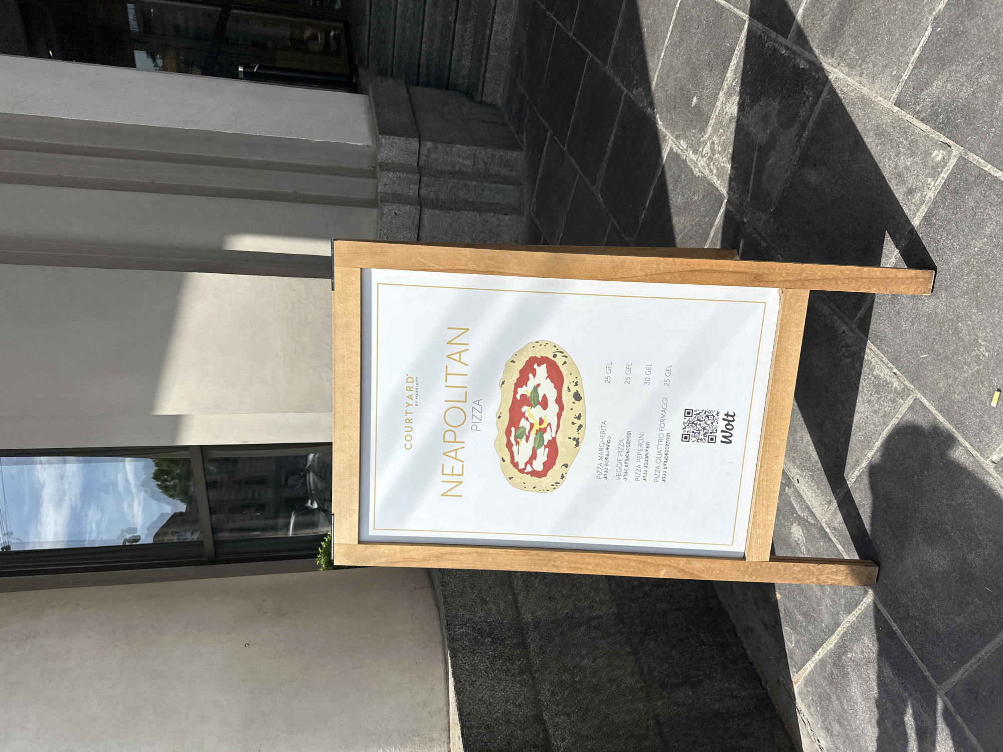 The image shows an advertising sign for "Neapolitan Pizza" at the Courtyard by Marriott, located in Tbilisi, Georgia. The sign is displayed on a wooden A-frame stand situated on a paved sidewalk next to the building. The menu includes Pizza Margherita, Veggie Pizza, Pizza Pepperoni, and Pizza Quattro Formaggi. The prices are listed in GEL (Georgian Lari). A QR code for Wolt, a food delivery service, is also present on the sign. The background includes a part of the building's facade with windows. The location is characterized by urban surroundings with stone pavement.