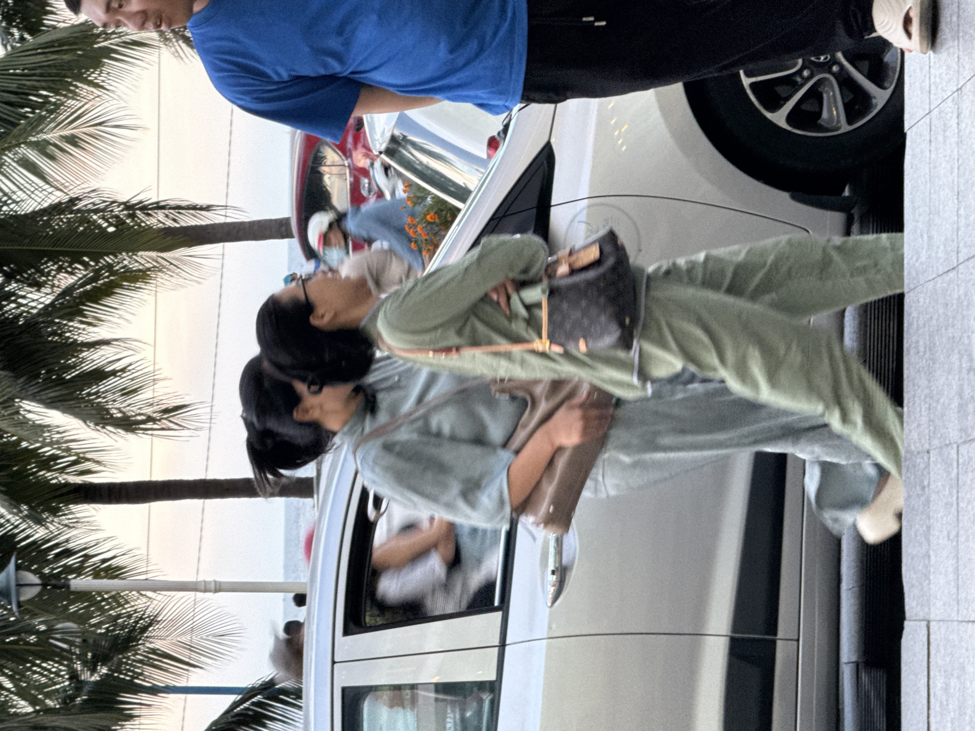 Two women are seen in front of a white car, with palm trees and the ocean in the background, suggesting a seaside location in Nha Trang, Vietnam. One woman, wearing a blue shirt, is standing and looking down. The other woman, dressed in light green clothing, appears to be leaning over to interact with the woman in the blue shirt. She is carrying a small, dark-colored handbag. The overall scene suggests a casual, outdoor interaction, possibly during the day given the bright sky.