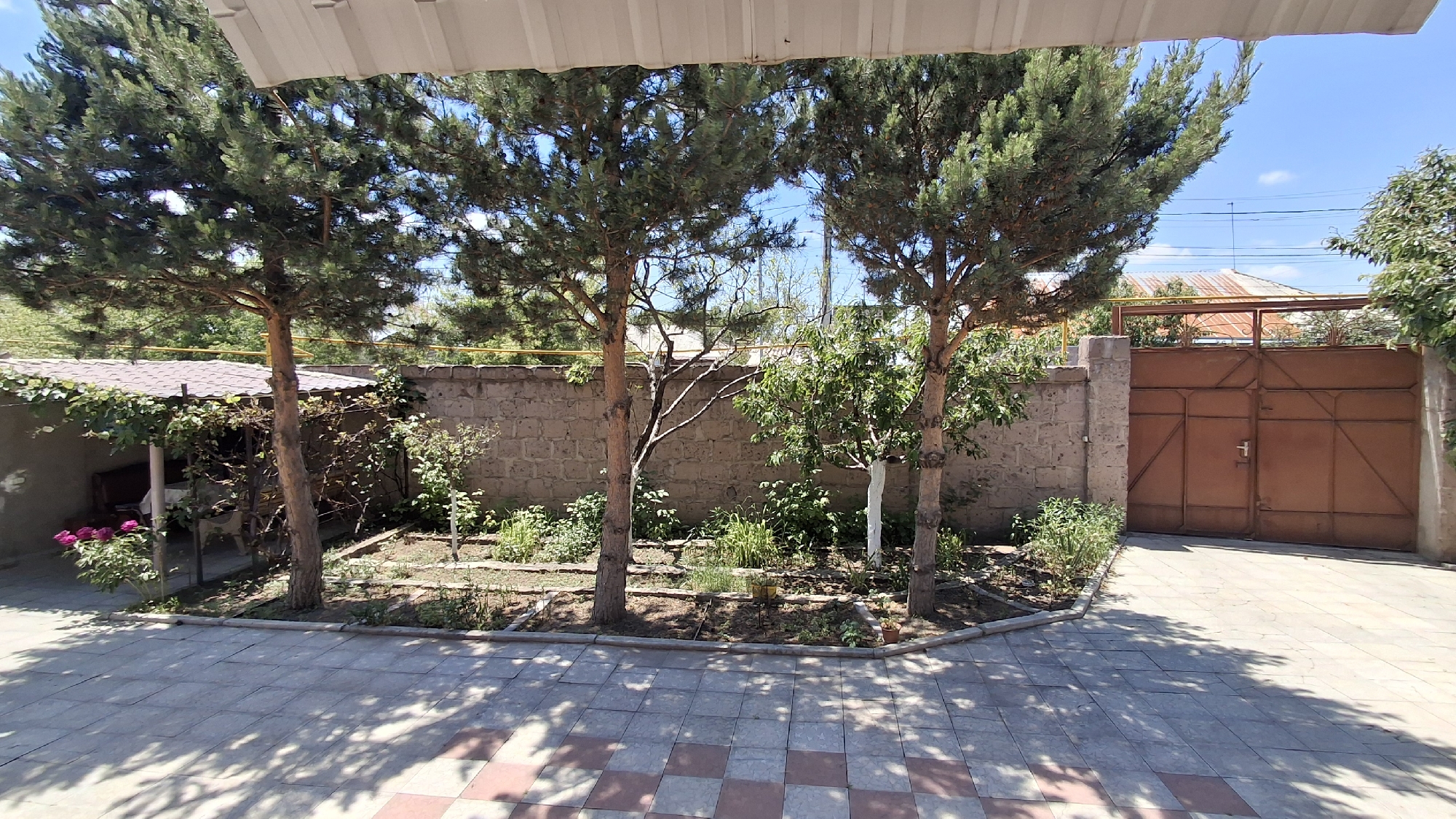 This is an outdoor medium shot of a courtyard with a paved walkway and a garden area. The walkway is made of square tiles in a checkerboard pattern. The garden is bordered by a low stone wall and contains various plants and small trees.
To the left, there's a covered structure with a brown roof, where some pink flowers are visible. Behind the garden is a rough stone wall. In the background, a large, rusty brown metal gate is visible, set within a stone wall. Beyond the gate, rooftops of houses can be seen, with one having a corrugated metal roof. The sky is blue with a few scattered clouds.
Three pine trees and two smaller deciduous trees are prominent in the scene, casting shadows on the walkway. The presence of a satellite dish and antenna on a distant rooftop suggests a residential area. The overall impression is a sunny day in a somewhat rustic, residential setting, possibly a backyard or entrance area. There is no visible text. The scene appears peaceful and quiet.