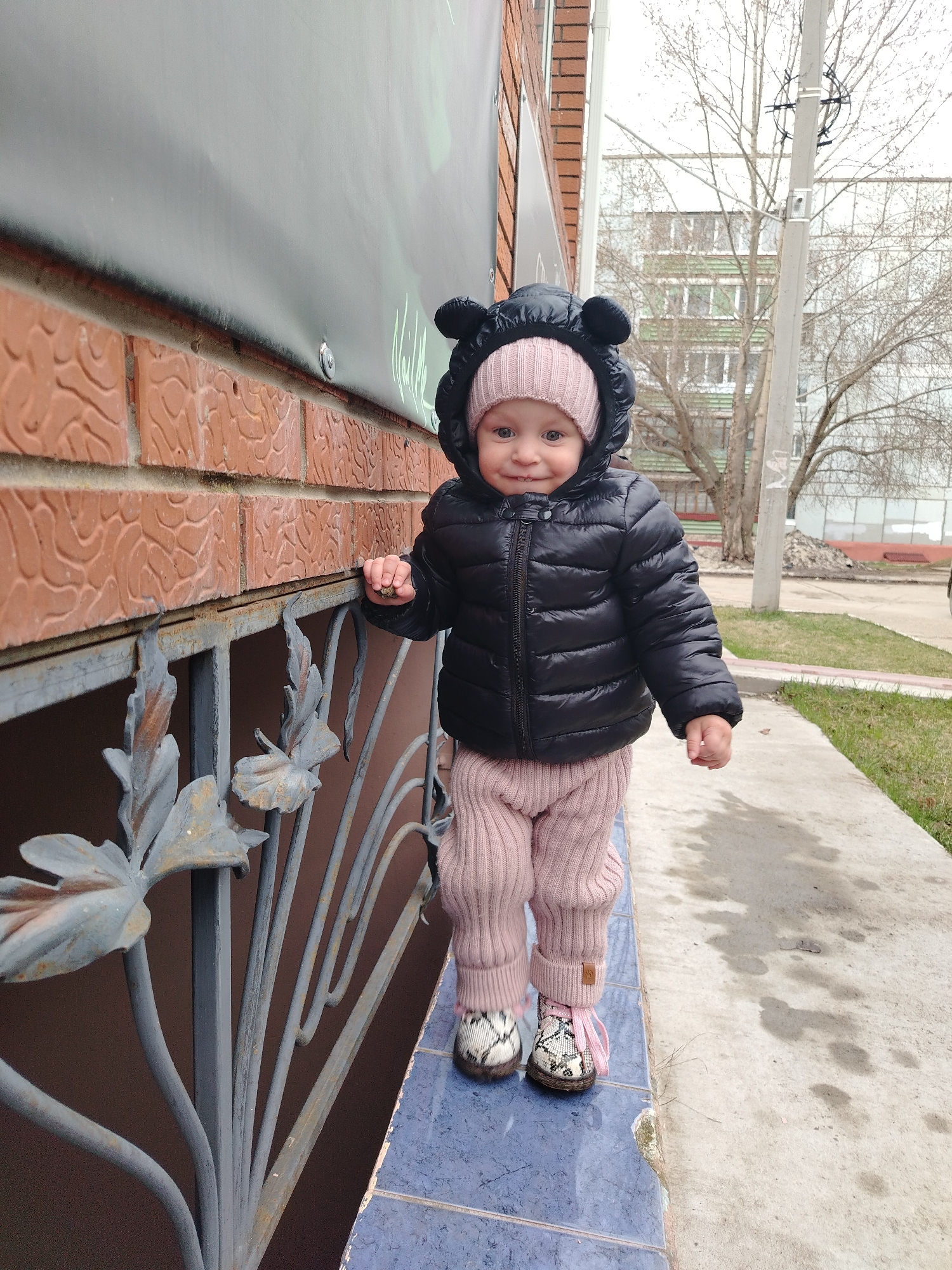 A cheerful toddler stands on a blue tiled ledge outdoors in Chaikovsky, Russia. The child, facing the camera, wears a black puffer jacket with a hood featuring prominent bear ears, a pink knitted beanie, pink ribbed pants, and light-colored shoes with a snakeskin pattern and pink laces. Their right hand is lightly placed near a decorative metal railing with leaf motifs, while their left hand is held slightly open. The child exhibits a slight smile and direct gaze, suggesting curiosity and happiness.
The setting appears to be an urban or suburban environment on a cool, overcast day, indicated by the bare trees in the background and the child's warm attire. Behind the child is a red brick wall with textured patterns, above which hangs a large, dark banner or sign with faint white stylized writing. To the right, a concrete sidewalk borders a strip of grass, leading to a utility pole and a distant, multi-story apartment building with green window frames. The tiled ledge the child is standing on separates the building facade from the ground level, with the ornate metal railing providing a decorative element below. The overall scene captures a moment of a child exploring their surroundings.