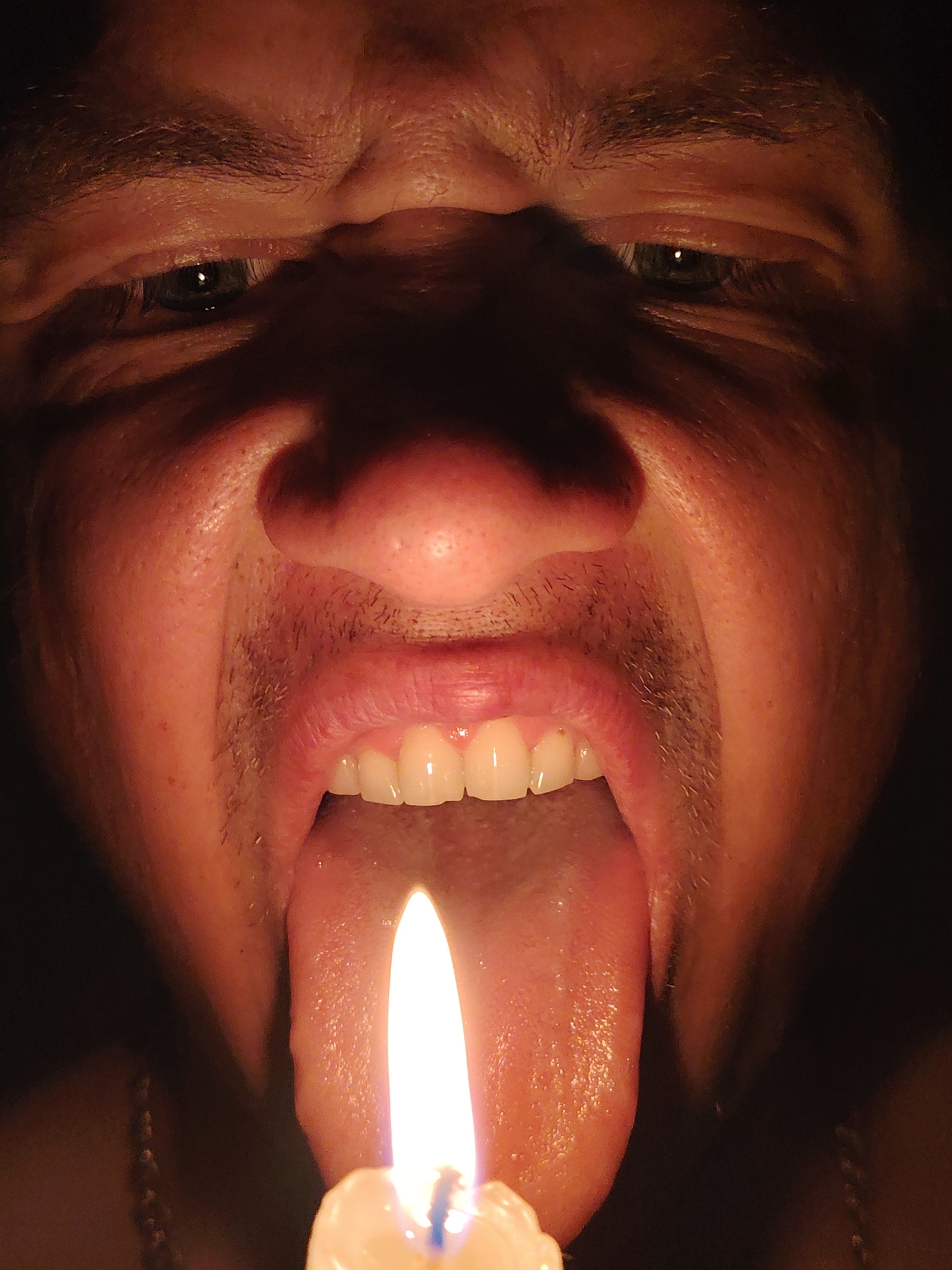 The image is a close-up, low-angle shot of a person's face with their mouth open and tongue extended. A lit candle is positioned in front of the tongue, with its flame illuminating the front of the tongue and the lower part of the mouth. The lighting is dramatic, with strong shadows obscuring much of the face and the background. The person's eyes are visible, looking towards the camera, and they appear to be dark. The skin texture is visible, and there is some facial hair. The background is entirely dark, suggesting an indoor setting with minimal ambient light. The image does not contain any specific geographical markers or context that would allow for identification of its location in Chaikovsky, Russia.