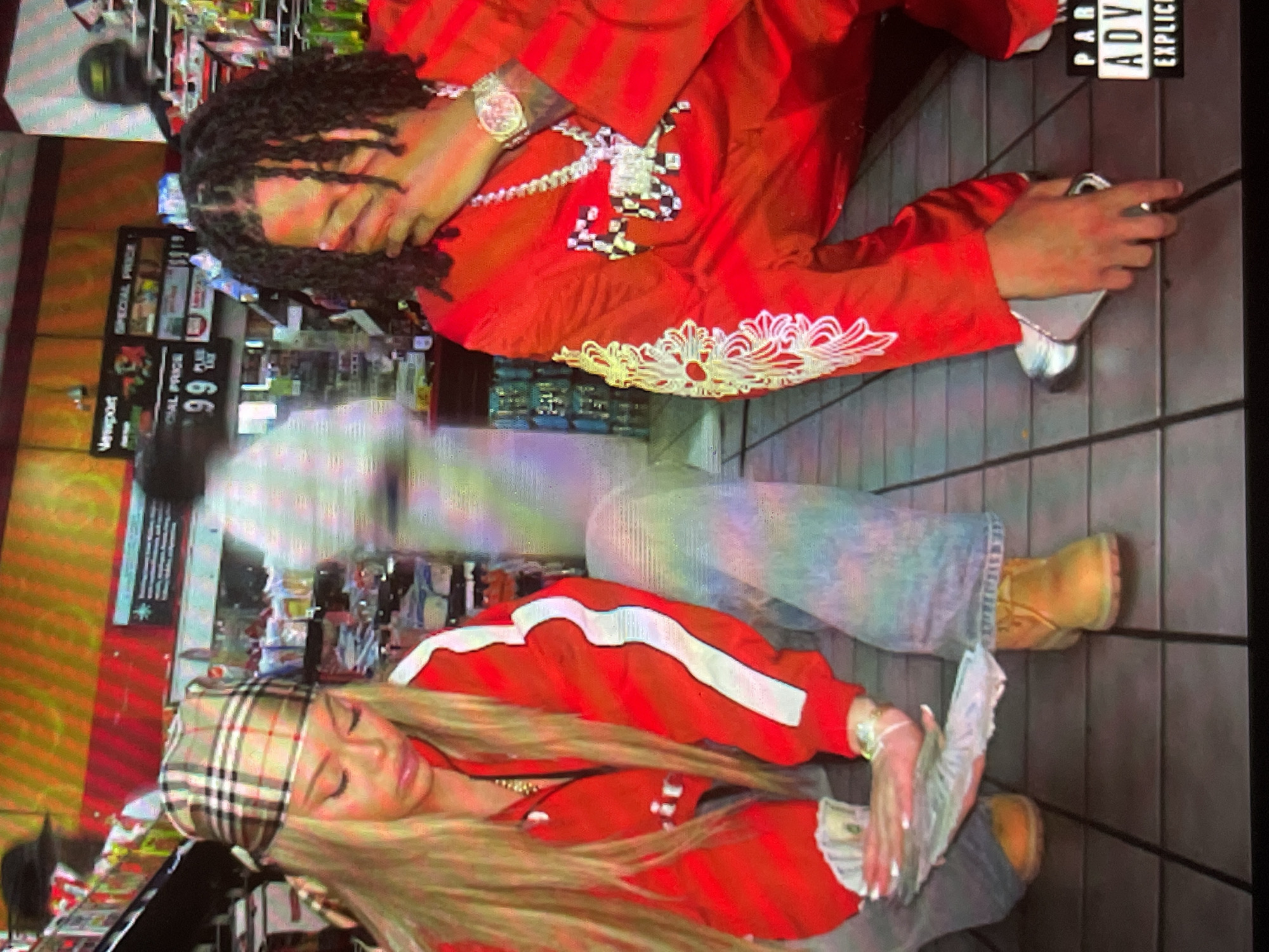 This vertical image depicts two individuals posing on the floor of what appears to be an indoor convenience store or gas station minimart. The background shows brightly lit shelves filled with various products, along with signs displaying "SPECIAL PRICE," "99" (likely cents), and "Vaporport."
In the upper part of the frame, a person with dark dreadlocks is positioned on their side. They are wearing a striking red long-sleeved shirt featuring a white and black checkerboard pattern across the chest and intricate white embroidery along the sleeve. This individual wears a large, ornate watch on their wrist and a prominent chain around their neck. Their eyes are closed or looking downwards, and they hold a silver-colored smartphone.
Below them, another person with long blonde hair is similarly posed on the floor. They are dressed in a red jacket with distinctive white stripes, blue jeans, and light brown boots. A plaid bandana, resembling a Burberry pattern, covers their forehead. This individual holds a fan of what appear to be US dollar bills in their hand, showcasing neatly manicured nails.
A third, heavily blurred figure is visible in the mid-ground, suggesting motion. On the right side of the image, a "PARENTAL ADVISORY EXPLICIT CONTENT" label, often associated with music, is partially visible. The overall scene suggests a stylized or staged photograph within a retail setting. The location context provided for this image is Sofia, Bulgaria.
