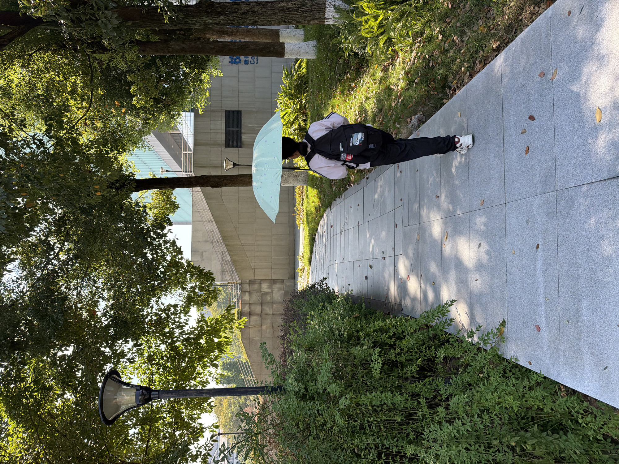 A person walks away from the camera along a paved path in a park-like setting. The person, viewed from the back, is wearing a white jacket with black trim, black pants, and white sneakers. They are carrying a black backpack and holding a light blue umbrella over their head. The path is made of large, light gray granite tiles and is bordered by lush green bushes on the left and grass and trees on the right. Sunlight filters through the leaves of the trees, casting dappled shadows on the path. In the background, there is a concrete structure with stairs and a modern building with a glass facade. The scene is set in the city of Jiangjin, China.