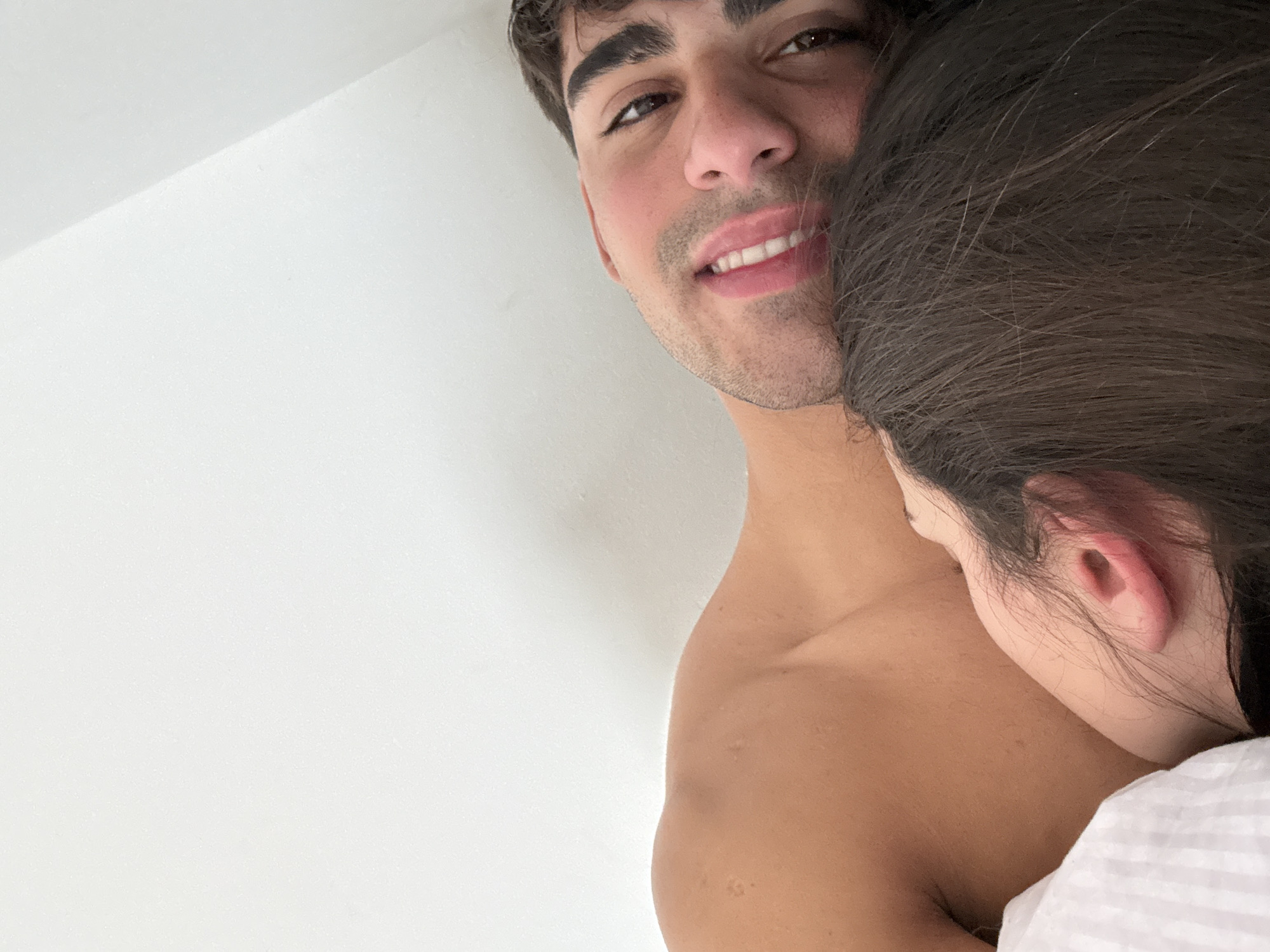 This is a close-up, selfie-style photograph taken indoors, likely in a bedroom. The image features a man and a woman in bed, positioned closely together. The man is partially visible, looking directly at the camera with a smile. His skin tone is tan, and he has dark hair and eyebrows. The woman is nestled against him, with her head resting on his shoulder. Only the side of her face and her ear are visible; she has long, dark hair. The background consists of a plain white wall, indicating an indoor setting. There are no distinct landmarks or features that would suggest a specific location within Avellaneda, Argentina. The overall scene conveys an intimate and relaxed atmosphere.