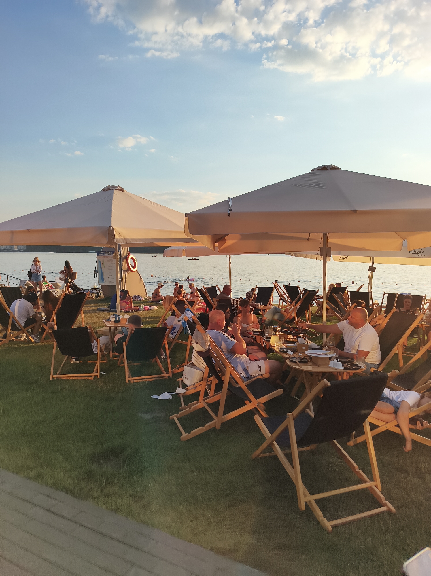 The image depicts an outdoor dining area on a grassy area next to a body of water, likely Ternopil Lake, in Ternopil, Ukraine. The scene is set during the late afternoon or early evening, with warm sunlight casting a golden glow. Several groups of people are seated at tables or in lounge chairs under large, light-colored umbrellas, suggesting a relaxed recreational atmosphere.
In the foreground, a group of individuals are seated at a round table, with food and drinks visible, indicating they are dining. Some are engaged in conversation, while others are looking at their plates or surroundings. Several wooden lounge chairs with black fabric seats are scattered throughout the scene, some occupied by people relaxing, others empty.
In the middle ground, more people are seen sitting at tables and chairs, some interacting with each other. The lake is visible behind them, with a few boats and people in the water, suggesting it is a popular spot for swimming or other water activities. The shoreline has a sandy or gravelly edge in some areas.
The background shows a wider expanse of the lake with more distant figures and structures. The sky is a clear blue with scattered white clouds, and the overall lighting suggests a pleasant, temperate climate. The presence of beach umbrellas and lounge chairs points to a beach or lakeside resort area. The overall impression is one of a lively and enjoyable outdoor gathering place.