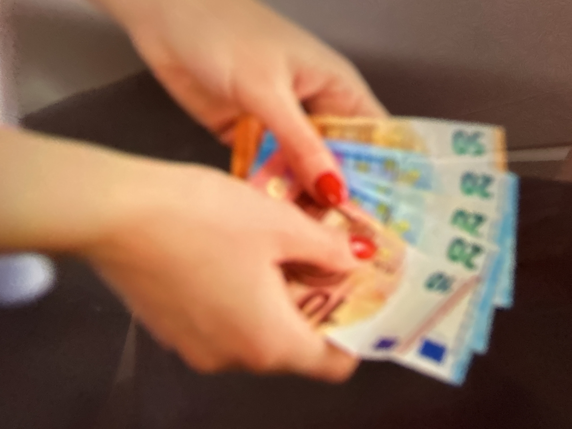 In the image, a person's hands are visible, holding a fan of euro banknotes. The hands are fair-skinned and adorned with red nail polish. The euro banknotes appear to be of different denominations, with visible numbers like "10", "20", and "50" on them, suggesting they are 10 euro, 20 euro, and 50 euro notes. The banknotes have a blue and yellow color scheme, typical of euro currency.
The scene is out of focus, making it difficult to discern the exact setting. However, the lighting suggests an indoor environment, possibly with artificial light. The background is dark and blurred, with a hint of a darker surface in the foreground, possibly a table. There are no clear indications of the time of day or weather conditions. The activity depicted is the handling or counting of money. No specific emotions are conveyed due to the lack of facial features and the overall blurriness of the image. No specific location cues are identifiable within the image itself, other than the currency being euro.