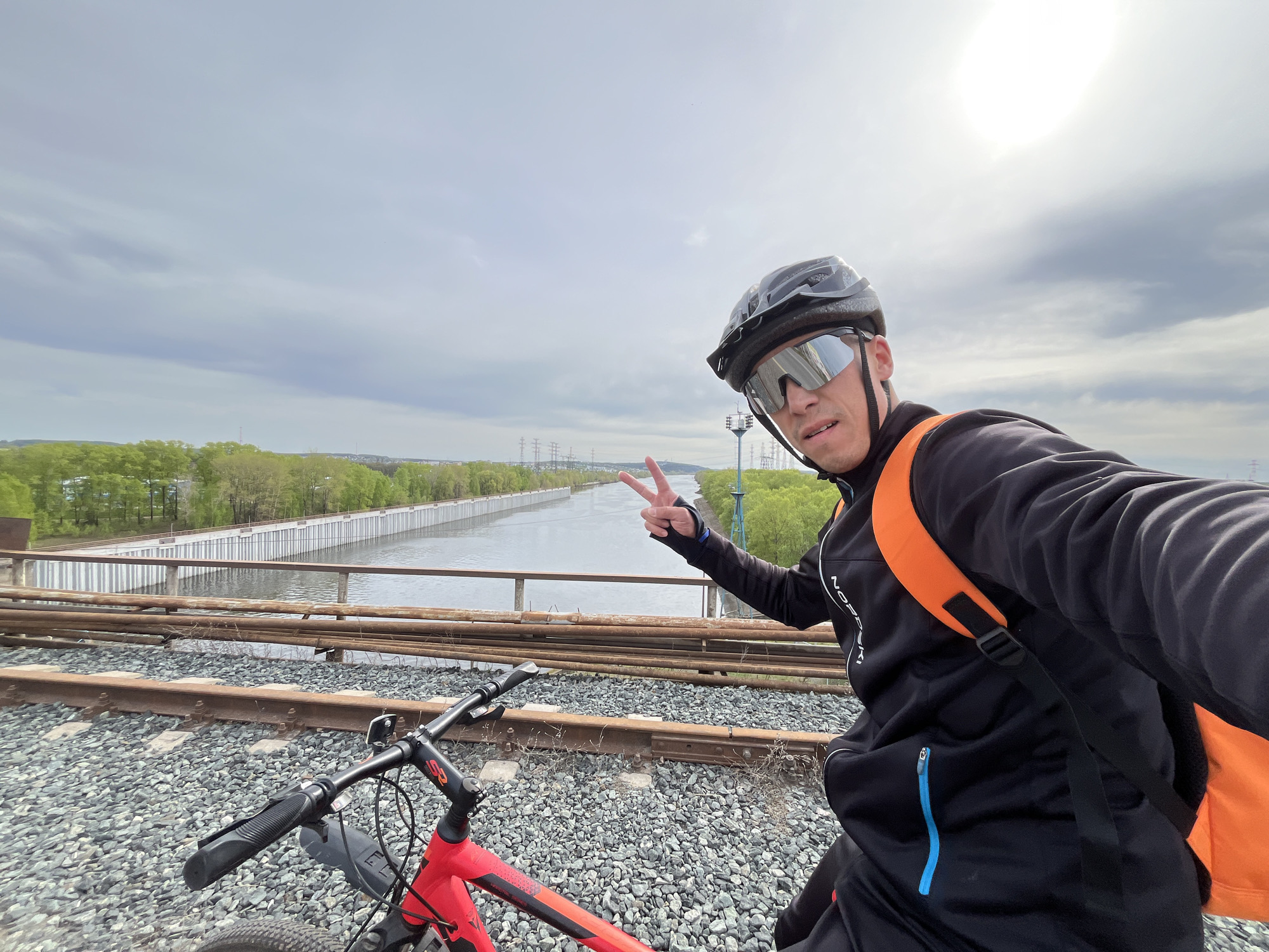 A man wearing a bicycle helmet and sunglasses is taking a selfie on a set of railroad tracks. He is on a red bicycle, and he is holding up two fingers in a peace sign. In the background, there is a canal, trees, and a cloudy sky. The power lines are in the background, and this is the location of Chaikovsky, Russia.
