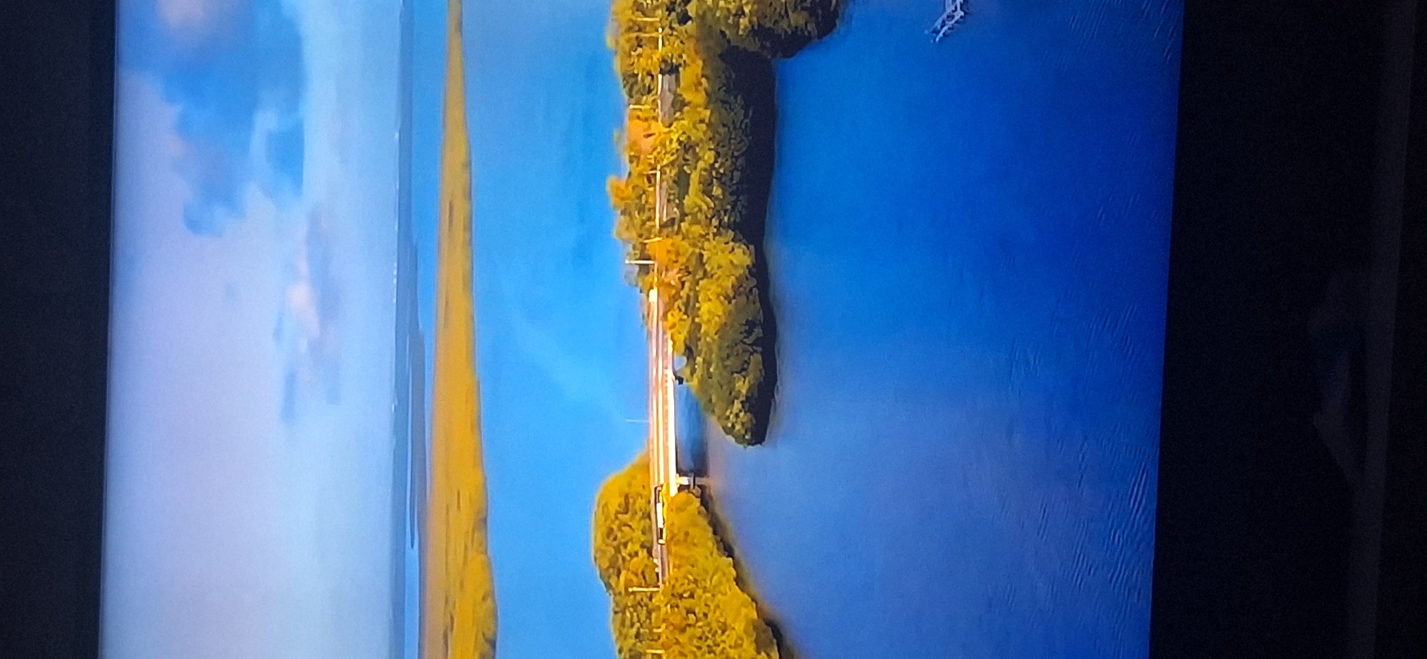 The image is an aerial view of a coastal landscape. In the foreground, a wide expanse of deep blue water stretches towards the horizon. A narrow channel of this water cuts through a densely vegetated island, with trees exhibiting a vibrant yellow-green hue, suggesting autumn foliage. A bridge spans this channel, connecting two parts of the island. Beyond this island, another body of calmer, lighter blue water is visible, bordered by a wide stretch of marshland in a golden-yellow color. In the far distance, a thin, hazy line likely represents a distant coastline or landmass. The sky above is a pale blue, with scattered white and gray clouds. The scene is serene and depicts a natural environment. The image does not provide any information about the city of Bila Tserkva, Ukraine.
