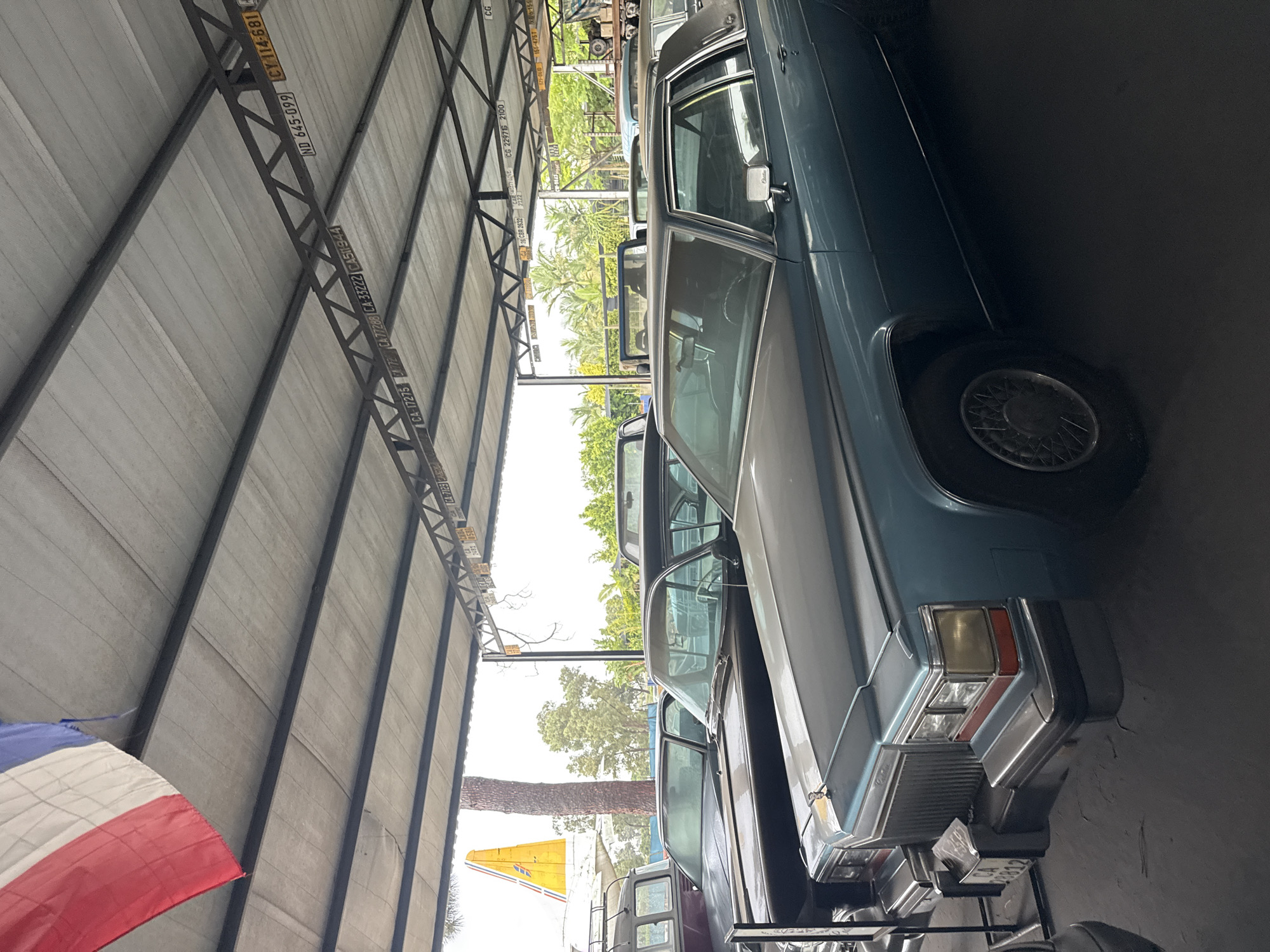 The image shows a row of vintage cars parked inside what appears to be a large garage or storage facility, possibly in Kraaifontein, South Africa, given the license plates. The cars are mostly light-colored sedans, partially covered, with one car taking center stage from a closer perspective. The structure above is made of steel beams supporting a roof. License plates are affixed to the beams. A flag is visible in the left corner. The environment suggests a collection or a storage area for classic cars.