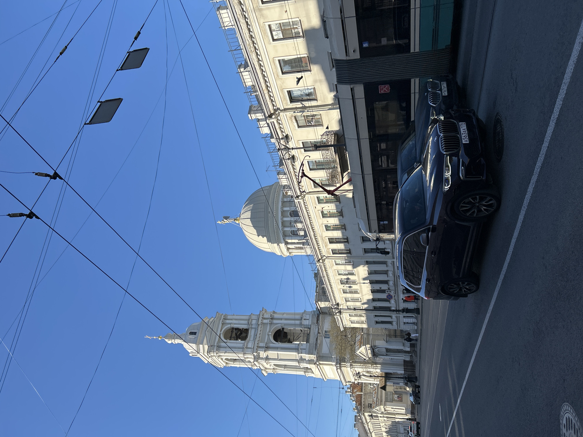 This image captures a bright, sunny urban street scene in Saint Petersburg, Russia.
The primary subjects include a striking white architectural complex dominated by an ornate bell tower with arched openings and a large dome, suggesting a church or cathedral, visible on the left side of the frame. Adjacent to it, a multi-story building with numerous windows and balconies lines the street.
On the right, an asphalt road features two dark-colored SUVs, identifiable as BMWs, one parked closer to the viewer and another further down the street. Behind them, part of a green and white tram or bus is visible. Overhead, a complex network of power lines stretches across a clear blue sky, with several rectangular panels attached to them, possibly sensors or electrical components. The road has clear white markings and a manhole cover is visible. A few indistinct figures can be seen walking on the distant sidewalk.
The time of day appears to be bright daylight, possibly morning or early afternoon, indicated by the strong light and clear, shadow-casting sun. The weather is sunny and clear.
Visible text includes Russian license plates on both BMWs: "A563AA 178" and "P161BP 178," with "178" being the region code for Saint Petersburg. A red "no entry" or "restricted access" sign and a blue sign with a white wheelchair symbol are visible on the side of the tram/bus.