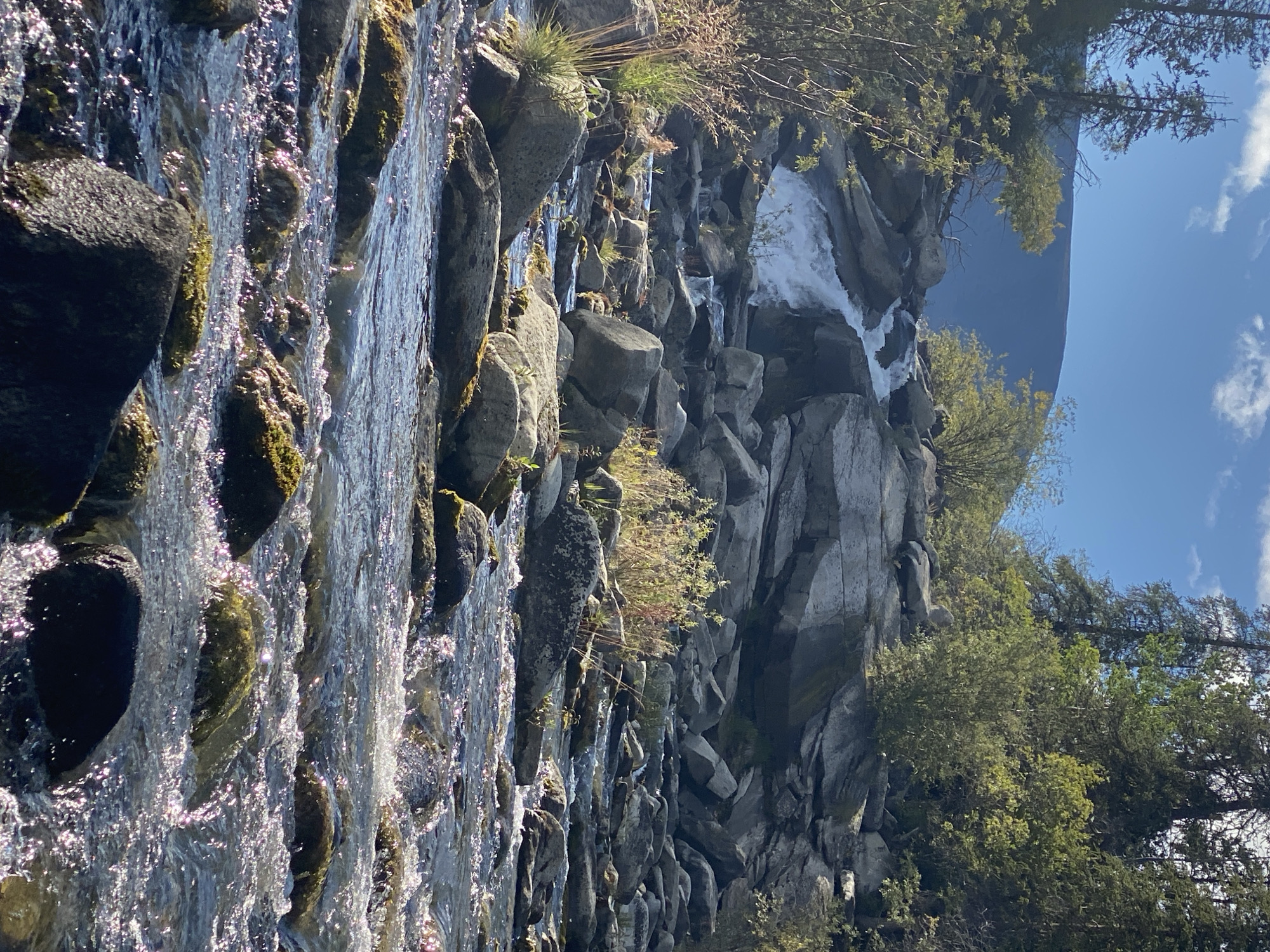 A natural landscape scene depicts a rocky river flowing downhill with a small waterfall in the distance. The foreground is dominated by the river's surface, which is filled with sunlight reflections and scattered rocks, some of which are covered in moss. The riverbed is also composed of numerous rocks of various sizes. In the middle ground, the river cascades over a series of rocks, forming a small waterfall. Surrounding the river are large, gray rocks and patches of dry, light-colored grass. The background features a dense forest of green trees, with a large, dark mountain visible behind the trees under a blue sky with scattered white clouds. The location is Yukon, Unorganized, Canada. There are no people or animals visible in the image.