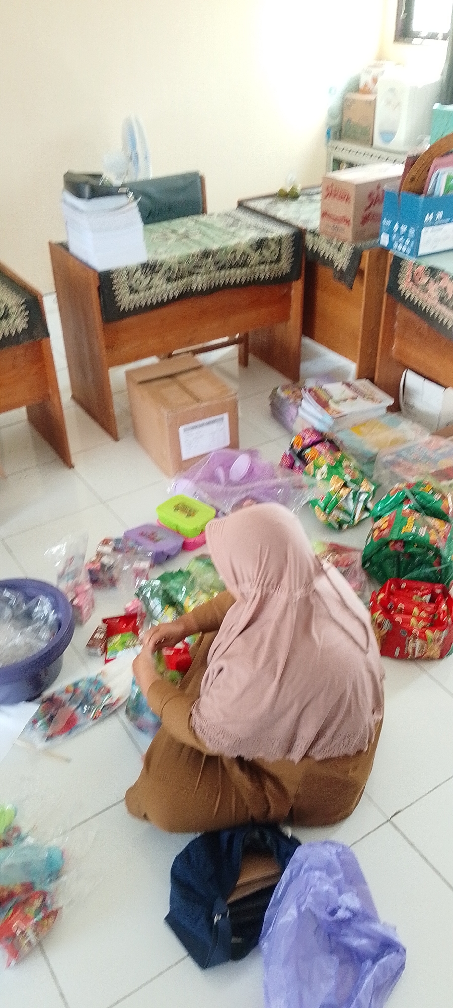 In Saring Sungai Binjai, Indonesia, a woman wearing a light brown hijab and a matching brown outfit sits on a tiled floor, occupied with preparing items. She is surrounded by numerous plastic-wrapped packages, some containing colorful snacks or toys, and several open plastic containers, one of which is a large purple bowl. In the background, two wooden benches are visible, each draped with a patterned cloth. On one bench, a stack of white papers and a dark object, possibly a bag, are present. Another wooden box is on the floor near the benches. The environment appears to be an indoor space with light-colored walls and a window or door frame visible in the upper right corner.