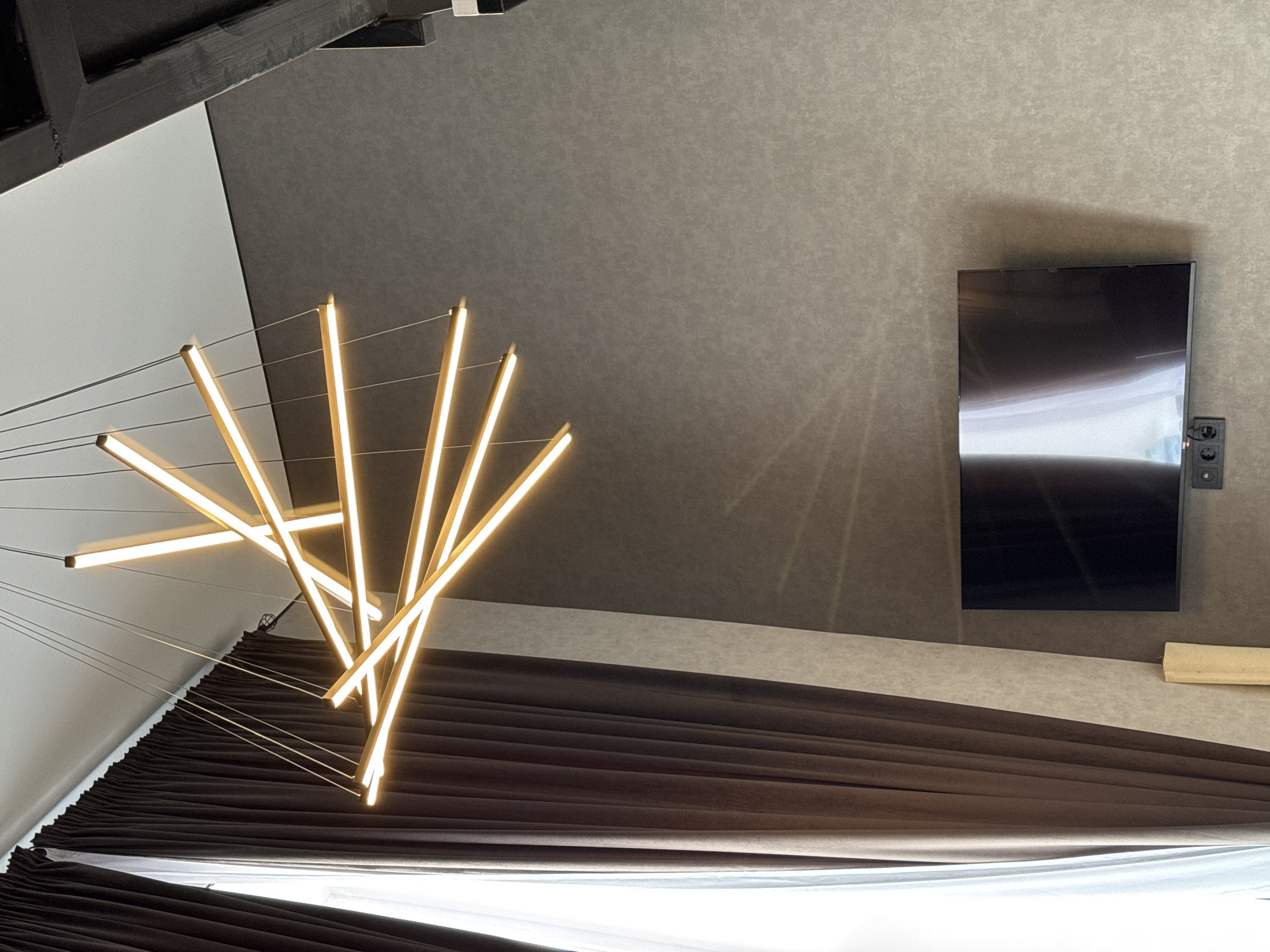 The image displays a modern interior space, captured from an upward-tilted angle. A striking, abstract chandelier, featuring multiple linear LED light bars emitting a warm, golden glow, dominates the left and central parts of the frame. These light bars are suspended by thin wires, creating a dynamic, crisscrossing pattern.
The background consists of a wall or ceiling covered in a textured, mottled grey-brown material. To the upper left, a section of a lighter, off-white or light grey surface is visible, along with a dark, possibly wooden or metal, beam structure.
On the right side of the grey-brown wall, a black flat-screen television is mounted. Adjacent to and below the TV, a black power outlet unit with visible plugs is attached. Further below the television, a light-colored, rectangular object, possibly made of wood, is affixed to the wall.
The lower portion of the image reveals heavy, dark brown or black curtains, drawn with numerous deep folds, suggesting a plush fabric. Below these curtains, a bright white fabric or surface, possibly a bedsheet or a lower curtain layer, is visible. The illumination primarily originates from the chandelier, implying an interior setting where artificial light is the main source, potentially indicating evening. No people or visible text are present. The scene conveys a sense of contemporary design and elegance.