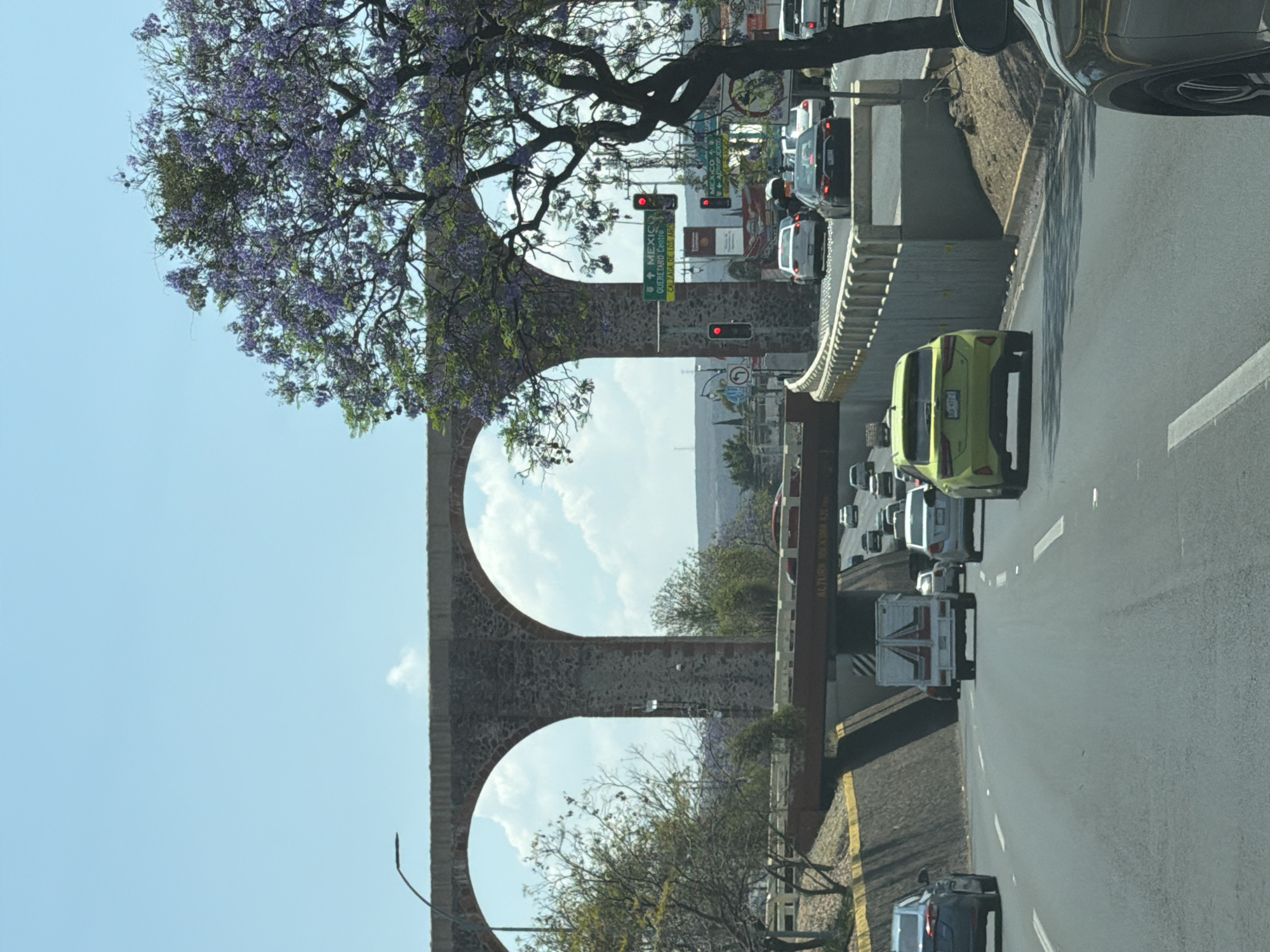 The image shows a traffic scene in an urban environment. A stone aqueduct with two prominent arches spans overhead. The aqueduct is situated on the left side of the frame, partially obscured by a flowering tree with purple blossoms in the upper section. Below the aqueduct, a road with traffic is visible, including a light green car and other vehicles. The traffic is dense, with multiple cars queued up. In the background, a cityscape and skyline are visible through the aqueduct's arches. Traffic lights and street signs are also present in the vicinity. The overall scene appears to be a busy intersection with a historical landmark. The location is likely Queretaro, Queretaro.