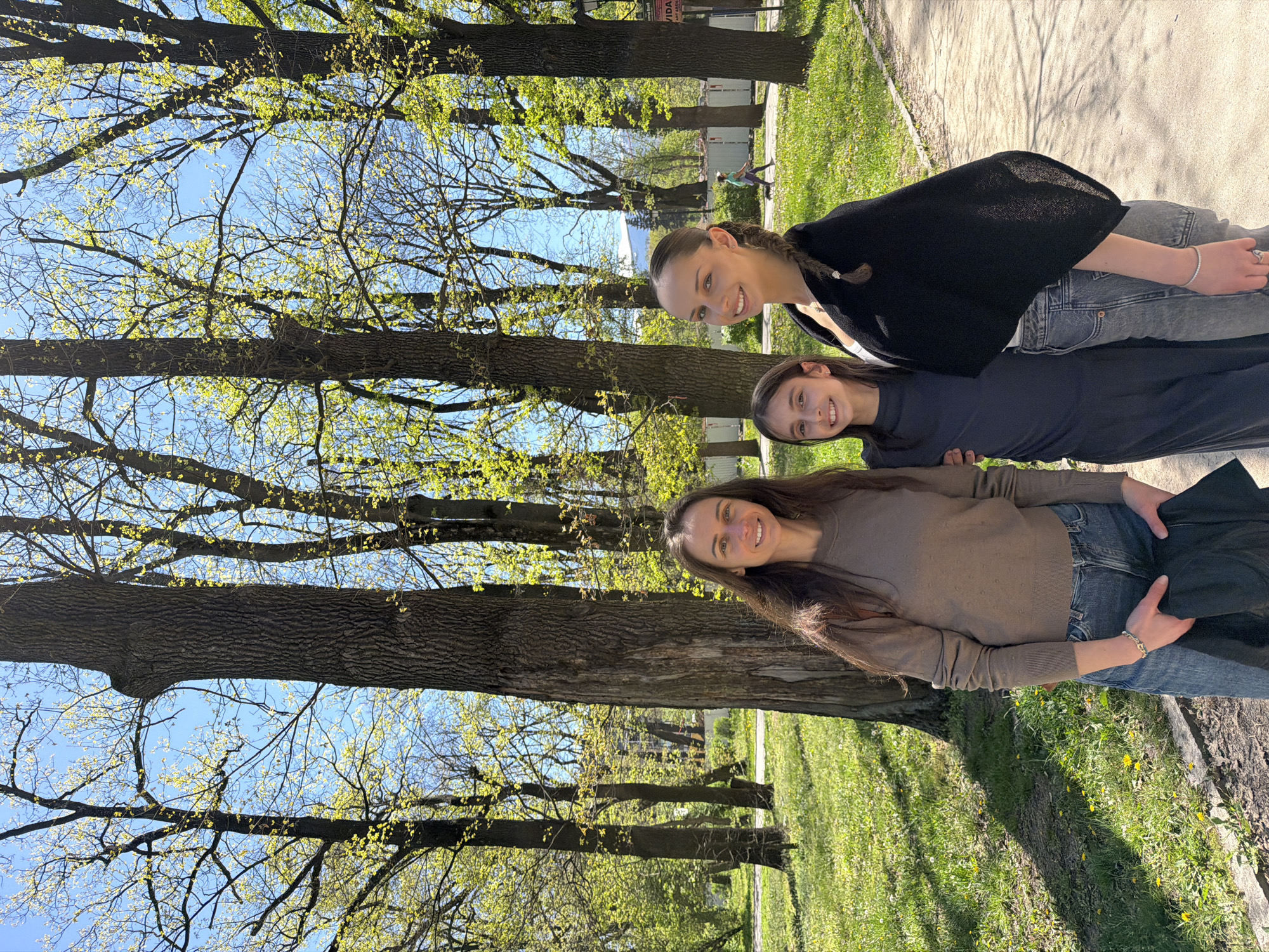 Three females of differing ages are positioned vertically in the image, likely in a park setting based on the presence of mature deciduous trees with spring foliage and grass, along a paved sidewalk. The setting may be the city of Sofia, Bulgaria, as suggested. The first woman is wearing a brown sweater and blue jeans; the second, a dark-colored dress; and the third, a black cardigan over a white top with gray jeans. They are all smiling and are facing the camera. The background consists of a tree-lined sidewalk, grass with yellow flowers, and a light blue sky visible through the tree branches. The overall light suggests daytime.