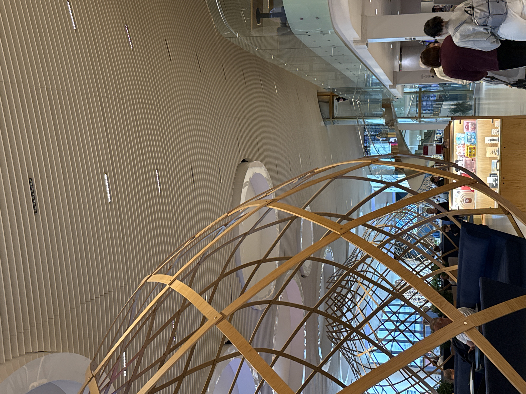 The image depicts the interior of a modern building, likely an airport or a large public space, with a focus on architectural design. In the foreground, a large, spherical wooden lattice structure dominates the view, its curved wooden beams forming an intricate geometric pattern. This structure appears to be a decorative element or part of a larger installation.
Beyond the wooden lattice, the space opens up to reveal a multi-level interior. A sleek, modern ceiling with linear lighting fixtures stretches across the upper part of the image. To the right, a balcony with a glass railing overlooks the lower level, with a spiral staircase and an elevator visible in the distance. People are seen walking and seated in this area, suggesting a functional public space.
In the lower left quadrant, blue upholstered seating is arranged, with several individuals seated and interacting with their surroundings. A display case with various small items is visible near the center of the image. The overall impression is one of contemporary design and spaciousness. The context of the city of Перепечино, Russia, is not discernible from the visual elements present in the image.