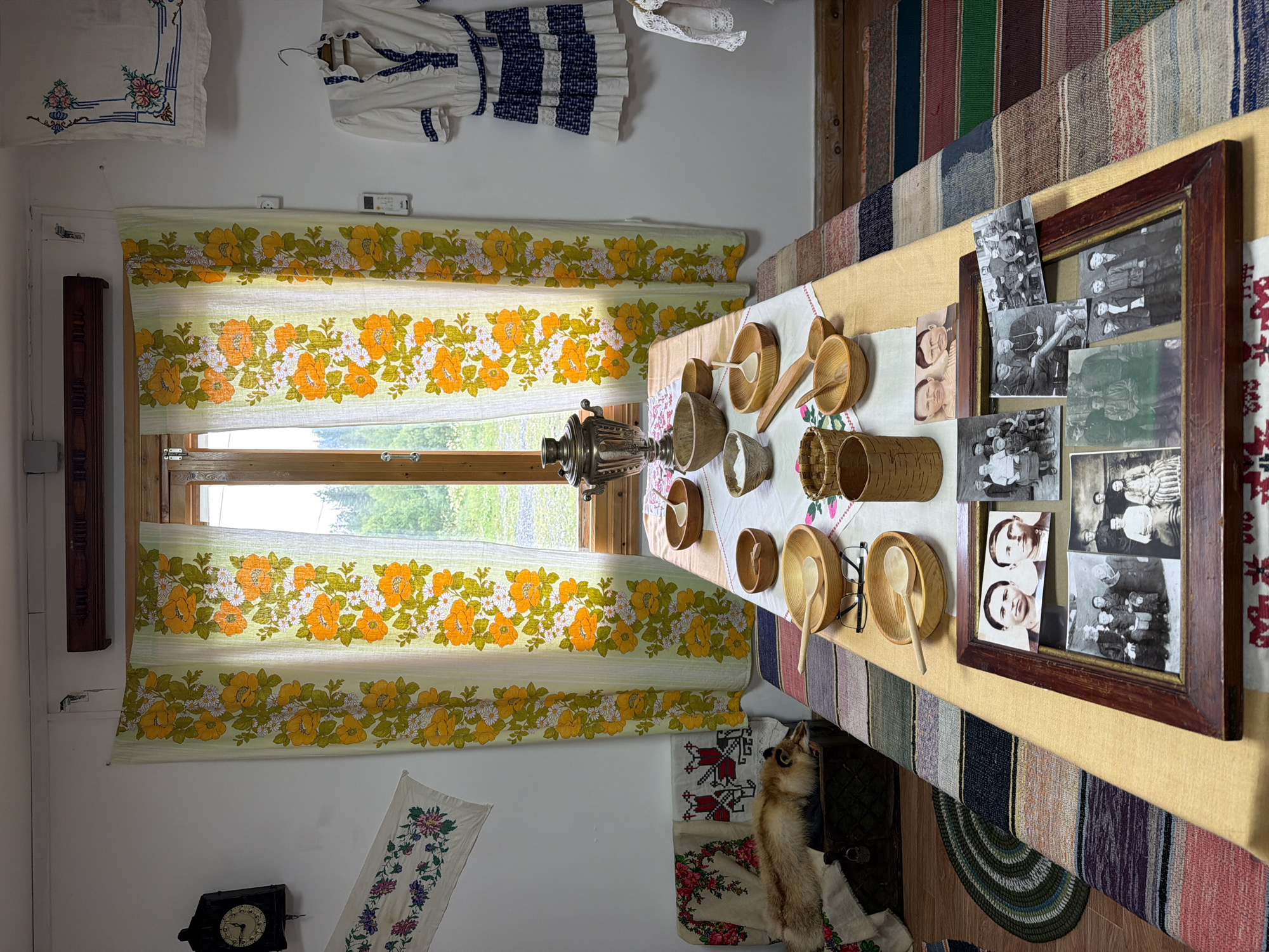 The image depicts an indoor scene, likely a room furnished in a traditional style. The room is decorated with floral curtains featuring orange and white flowers on a light green background, and several pieces of embroidered fabric. On the left wall, a black pendulum clock hangs, and below it, an embroidered runner is displayed. To the right of the window, a white and blue dress with ruffled detailing is hung on a hanger.
In the center of the room, a long table is covered with a light-colored tablecloth. The table is set with various wooden bowls, some containing spoons, and a samovar. There are also several framed photographs on the table, showcasing groups of people in vintage attire, as well as individual portraits.
On the left side of the room, near the window, a fox pelt is laid out on a chest, accompanied by embroidered cloths. The floor is covered with a striped rug. The overall impression is that of a culturally preserved space, possibly a museum exhibit or a well-maintained traditional home. The location is identified as Ust'-Pyzas, Russia, suggesting a cultural context related to that region.