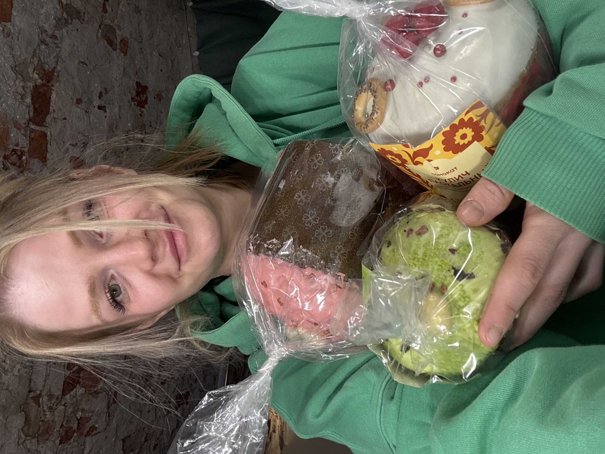 A young woman with light blonde hair and fair skin, wearing a green hooded sweatshirt, is holding several wrapped Easter cakes (Kulich). One cake is white with red decorative elements and a yellow-orange label with Russian text. Another is brown with a pink top and floral patterns. A third is green with dark red specks. The background includes a brick wall. The scene potentially takes place in Saint Petersburg, Russia, based on the style of Easter cakes.