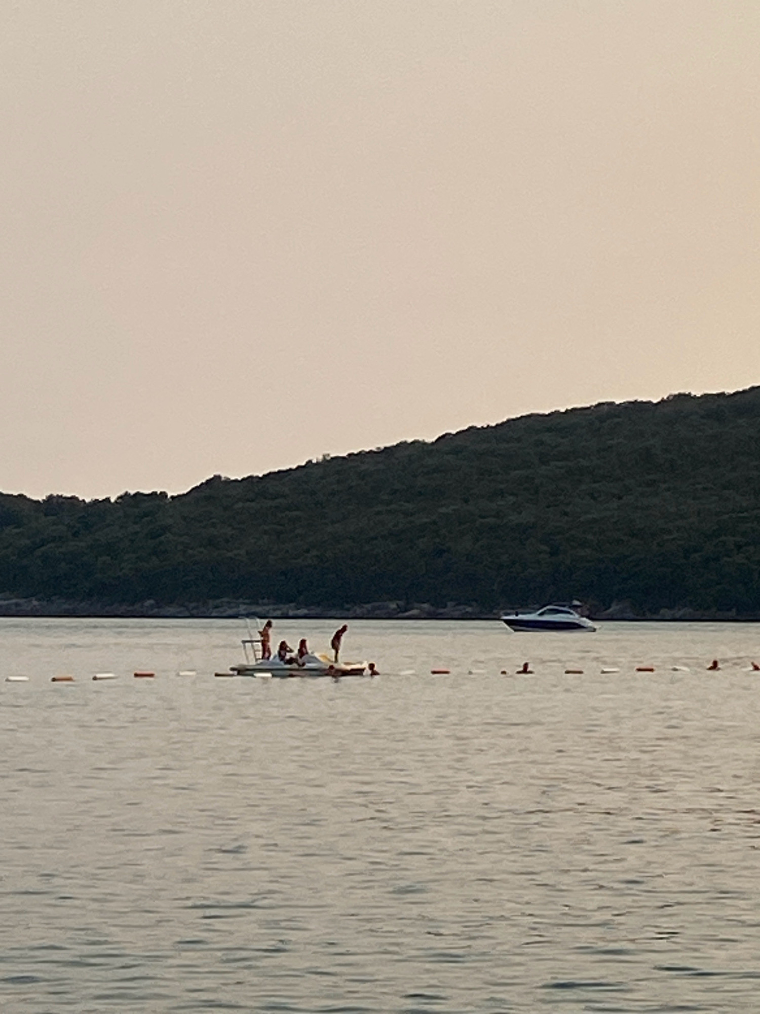 The image captures a serene water scene in Prijevor, Montenegro, dominated by a calm sea and a lush, dark green forested hill that slopes down towards the water's edge. The sky is a soft, pale gradient of pink and beige, suggesting either dawn or dusk. In the middle ground, a group of people are gathered on a small, floating platform or raft. Several individuals are standing, and some appear to be interacting or possibly preparing to jump into the water. A white and blue motorboat is visible in the distance, cruising across the water parallel to the shoreline. A rope with alternating white and orange buoys stretches across the foreground, marking a boundary or a designated swimming area. The overall atmosphere is peaceful and recreational.