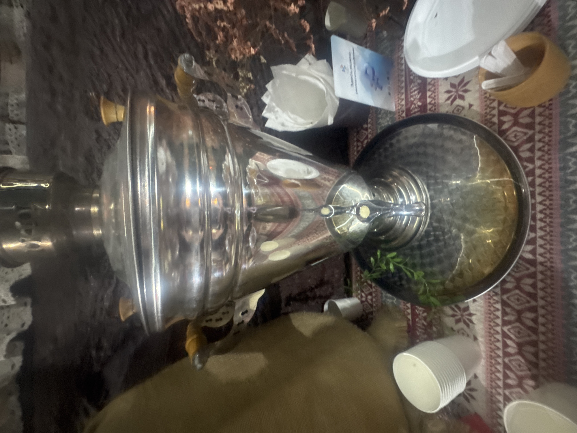 This media file features a close-up, rotated view of a shiny, metallic samovar, a traditional Russian teapot, placed on a patterned rug. The samovar is the central focus, with its reflective surface showing distorted images of its surroundings. The rug beneath has a reddish-brown and white geometric pattern.
To the left of the samovar, a dark, textured surface, possibly a rough wall or a dark fabric, is visible. To the right, a white paper cup is positioned on the rug. Further right, a flat, silver tray is partly visible, containing some greenery, possibly artificial leaves. Above the samovar, a white paper object, possibly a folded napkin, is placed next to a small white card with some text and a blue symbol. In the background, a white oval object, likely a plate or bowl, and a small woven basket containing white material are also present.
The lighting suggests an indoor setting, possibly with artificial light, as there are reflections and highlights on the metal surfaces. The orientation of the image suggests it was taken in Vietnam, specifically in Bac Nha Trang Ward, as per the location context. No specific activity or event is clearly depicted, but the presence of the samovar and cups might imply a gathering or a tea-drinking occasion. The overall mood is neutral, focusing on the objects and their arrangement. There is some visible text on the white card, but it is illegible in this image.