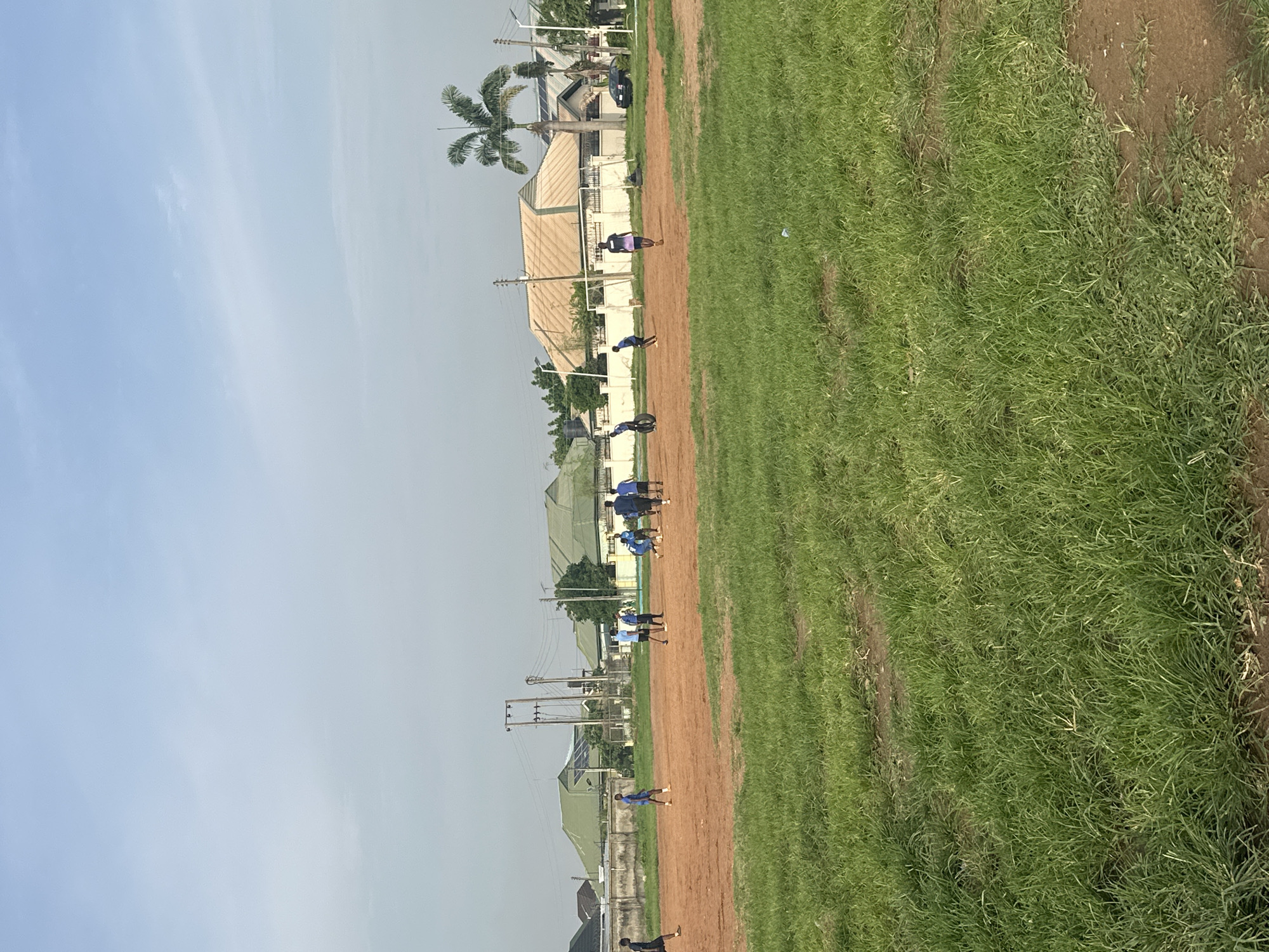 The image depicts an outdoor scene on a dirt and grass field, likely a football pitch, in Abuja, Nigeria. A group of young men are engaged in a football game. The environment is characteristic of a local park or sports ground, with buildings and vegetation visible in the background. The sky is partly cloudy. The presence of a palm tree and the architectural style of the buildings suggest a Nigerian setting, consistent with the city of Abuja. The field is primarily a reddish-brown dirt surface with patches of green grass, typical of many sports fields in the region. The people are dressed in athletic attire, and their postures indicate active participation in the game. The overall atmosphere appears to be one of casual sports activity in a community setting.
