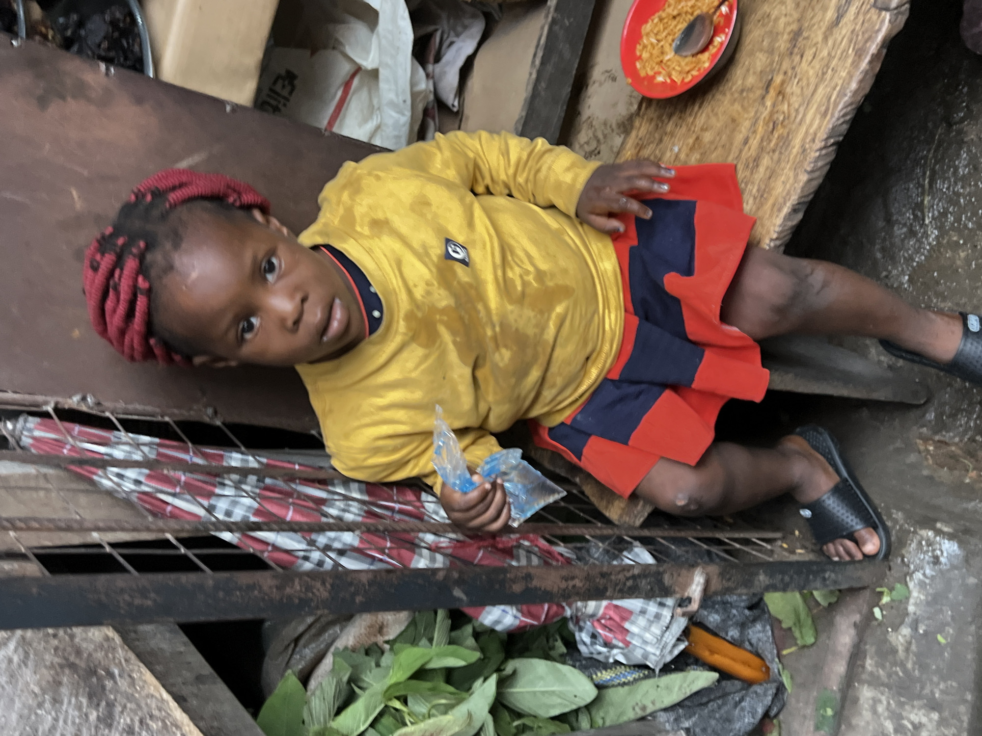 A young Black child, possibly female, with red braided hair sits on a wooden bench in what appears to be an outdoor setting. The child wears a yellow long-sleeved shirt and a red and navy blue skirt. A clear plastic bag containing a clear liquid is held in the child's right hand. To the child's right, a red bowl contains food and a spoon. In the background, there is a metal gate, some green foliage, a folded umbrella with a red, white, and blue checkered pattern, and various bags and boxes. The ground is wet and appears to be made of concrete. The overall scene suggests a humble environment, possibly a market or residential area in Nkpor, Nigeria.