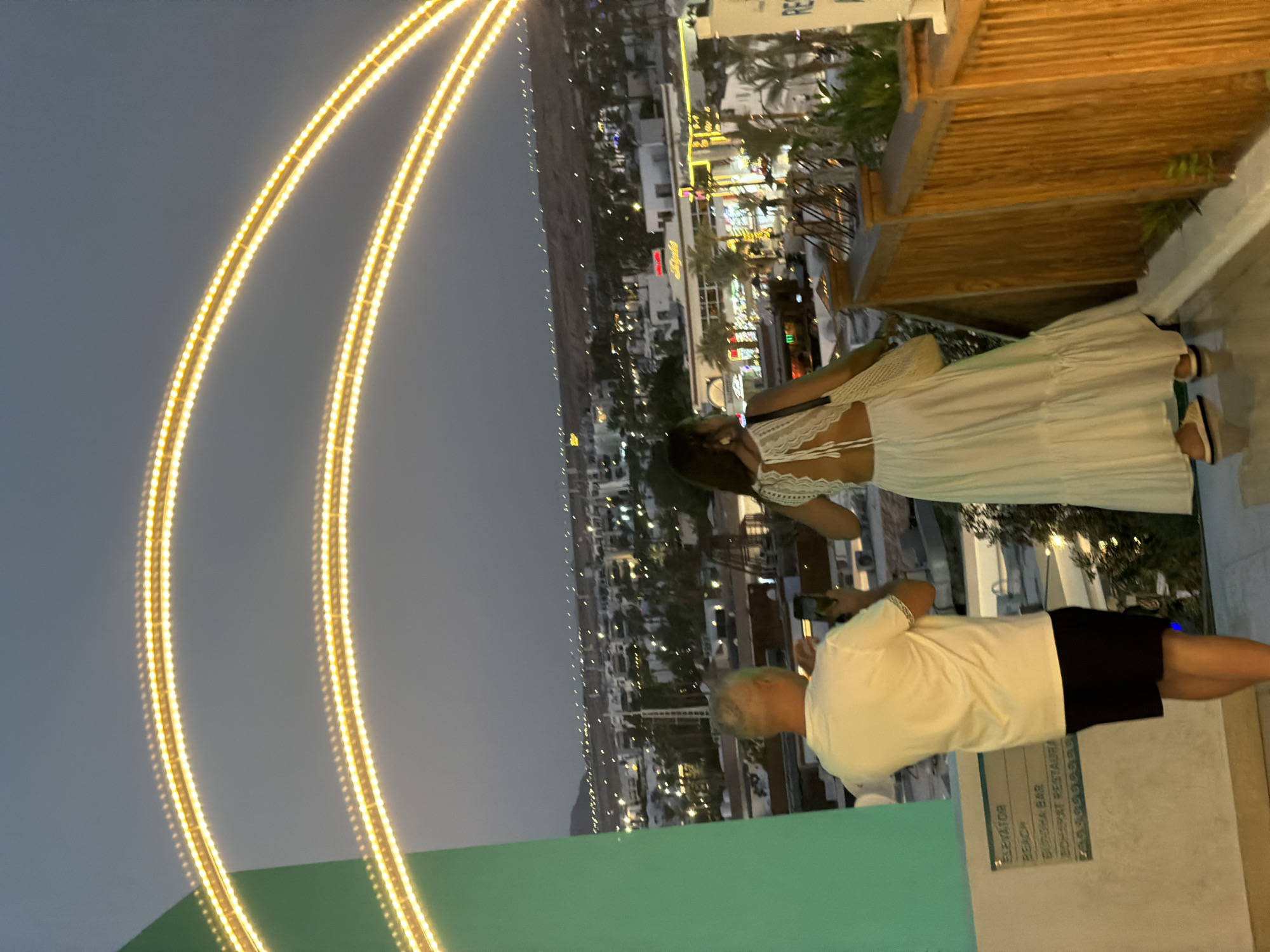 The image captures a twilight scene in Sharm El Sheikh, Egypt, overlooking a cityscape dotted with lights. In the foreground, two individuals are standing on an elevated platform, likely a balcony or terrace. A man, viewed from behind, is wearing a white t-shirt and black shorts, holding a smartphone and appearing to take a photo. Beside him, a woman in a flowing white dress with an open back is also seen from behind, carrying a woven bag over her shoulder.
The background reveals a sprawling urban landscape with numerous buildings and illuminated signs, stretching towards a dimly lit mountainous horizon. Palm trees are visible, suggesting a tropical or sub-tropical environment. Prominently featured in the upper portion of the image are two large, curved arches made of warm-toned LED lights, adding a decorative element to the scene. A green structure is partially visible on the left side of the frame. To the right, a wooden railing with vertical slats delineates the edge of the viewing area. A sign with text, partially obscured, is mounted on the green structure to the left, indicating "ELEVATOR", "BEACH", "BUKCHA BAR", and "BOHARAT RESTAURANT". The overall atmosphere is serene, with the city lights beginning to twinkle as dusk settles.