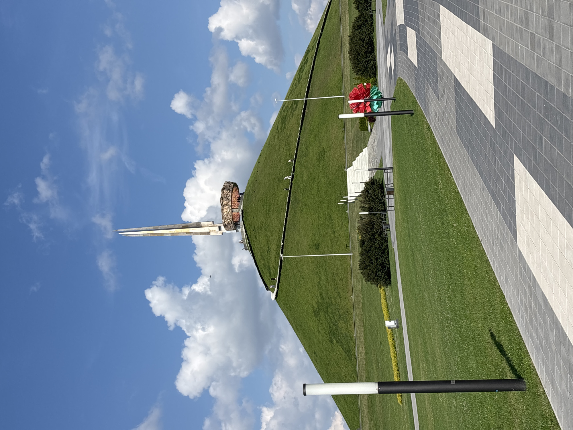 This is a medium-angle, eye-level, outdoor, daytime photograph featuring the Kurgan of Glory, a war memorial located in the Ozyaritska-Slabadski rural council, Belarus. The memorial is situated atop a large, green, grass-covered mound. A tall, slender obelisk, composed of metallic elements in shades of gold and beige, rises from the summit of the mound. Surrounding the base of the obelisk is a circular bronze frieze depicting faces and text. A winding pathway with steps ascends the side of the mound, leading towards the memorial. Several individuals are visible walking on this path. In the foreground, a paved walkway with a pattern of grey and white tiles curves towards the right side of the frame. Two modern lampposts are positioned along the walkway, one to the left and another to the right, with a decorative red and green floral wreath placed near the right lamppost. The environment is characterized by a bright blue sky dotted with fluffy white clouds, and well-maintained green grass covering the landscape.