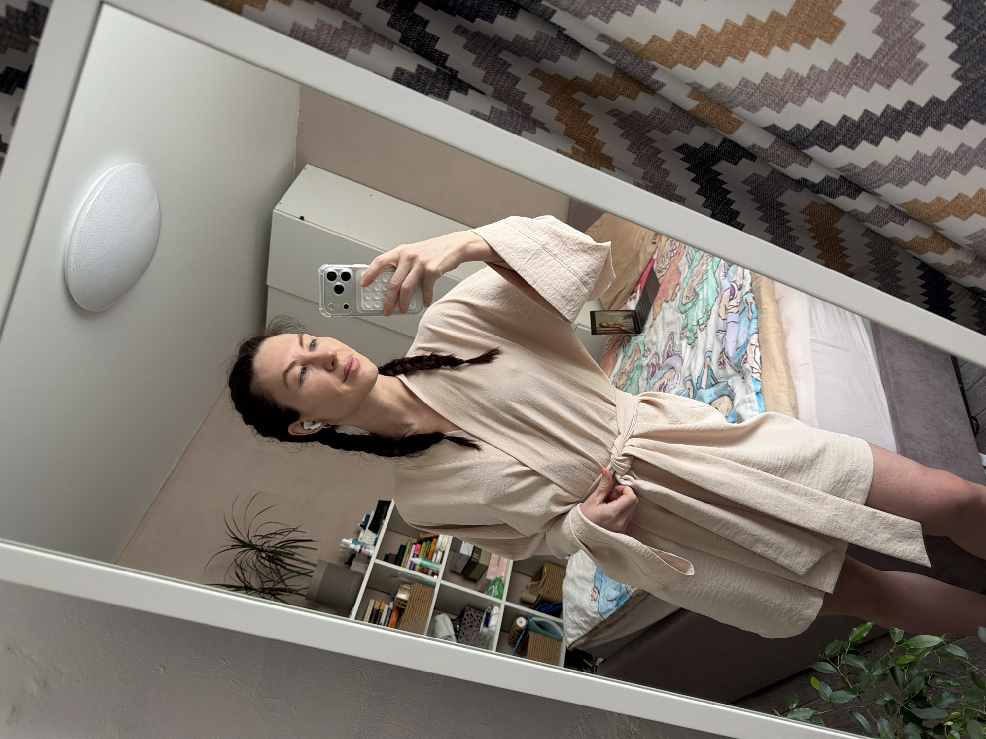 This image depicts a female individual taking a mirror selfie in a residential interior located in Perm, Russia. The subject, a woman with dark hair braided into two pigtails, is clad in a light beige, textured robe tied at the waist. She wears white wireless earbuds in both ears. Her right hand holds a silver-colored smartphone, equipped with a clear, possibly ridged, case, angled to capture her reflection in a large, white-framed mirror. Her head is tilted slightly, and her gaze is directed towards her right.
The reflected background reveals a room with light-colored walls and a white ceiling, from which an oval-shaped, flush-mount light fixture is suspended. To the subject's left, a white shelving unit is visible, filled with books, baskets, and various bottles or containers. Above the shelving unit, a potted plant with green foliage sits in a grey pot. A bed with a grey upholstered headboard or frame is partially visible, covered by a multi-colored duvet featuring animated or illustrated characters. A laptop with an illuminated screen displaying a person's face rests on the bed. To the subject's right, a section of a white cabinet or wardrobe is visible, adjacent to curtains displaying a zig-zag pattern in white, black, grey, and brown/gold hues. A portion of another green plant is visible in the lower left foreground of the reflected scene.