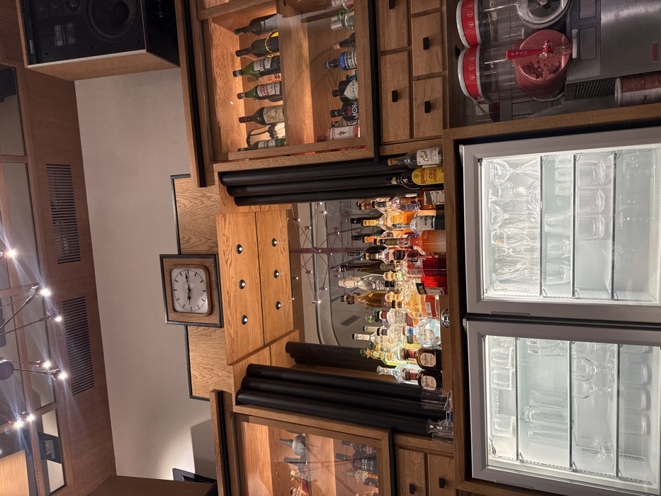 The image provides a detailed view of a bar area within The Loop Cafe in Yerevan, Armenia. The perspective is a medium-shot, looking upwards at the central and upper sections of a wooden bar structure.
At the center, a large rectangular mirror is flanked by dark, vertical cylindrical decorative elements. This mirror reflects a portion of the ceiling and a large, intricate, light-colored structural element, consistent with a Ferris wheel, located further within the establishment. Positioned above the mirror, embedded within the wooden paneling, is a square wall clock with a white face and black hands, indicating approximately 2:50.
The bar surface directly below the mirror is populated with a wide array of liquor bottles, including various tequilas, rums, gins, whiskies, and liqueurs such as Aperol, Kahlua, and Campari. Many bottles are branded and recognizable. To the left and right of the central mirror, integrated into the light-colored wooden cabinetry, are glass-fronted display cases, illuminated by recessed spotlights. These cases showcase additional bottles of spirits and wines, with smaller, ornate bottles visible on the left and a mix of wine bottles and various gin bottles on the right. Below these display cases are rows of wooden drawers with dark handles.
The lower portion of the image features two under-counter, glass-door refrigerators, internally lit and filled with a diverse collection of clean, empty glassware, including wine glasses, tumblers, and highball glasses, neatly arranged on shelves. To the far right, on the counter, are two commercial beverage dispensers with red lids, one appearing to contain a reddish slush. A large, dark-colored speaker cabinet is mounted high on the right wall, above the display cases. The ceiling, visible in the upper part of the image, is composed of wooden panels and features recessed ventilation grilles and modern, multi-armed chandelier-style light fixtures with exposed bulbs. The entire scene is brightly illuminated, and no individuals are present.