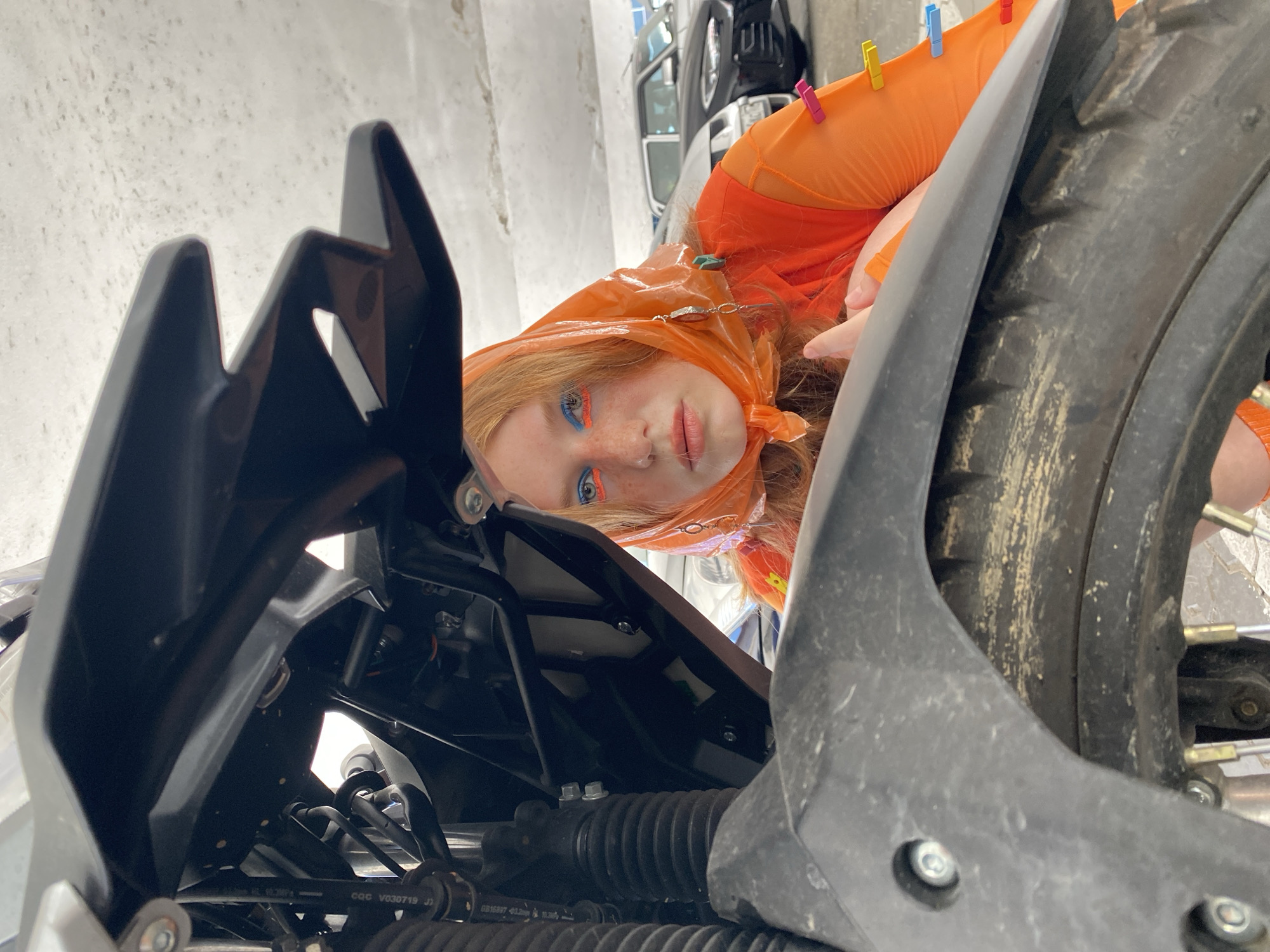 A young woman with auburn hair and bright blue and orange eyeshadow is peeking out from under a black motorcycle fairing. She is wearing an orange headscarf and an orange mesh top, which is adorned with several colorful clothespins. The motorcycle appears to be in a parking garage, with cars visible in the background. The concrete ceiling and walls of the garage are visible. The overall lighting suggests an indoor, possibly dimly lit environment.
