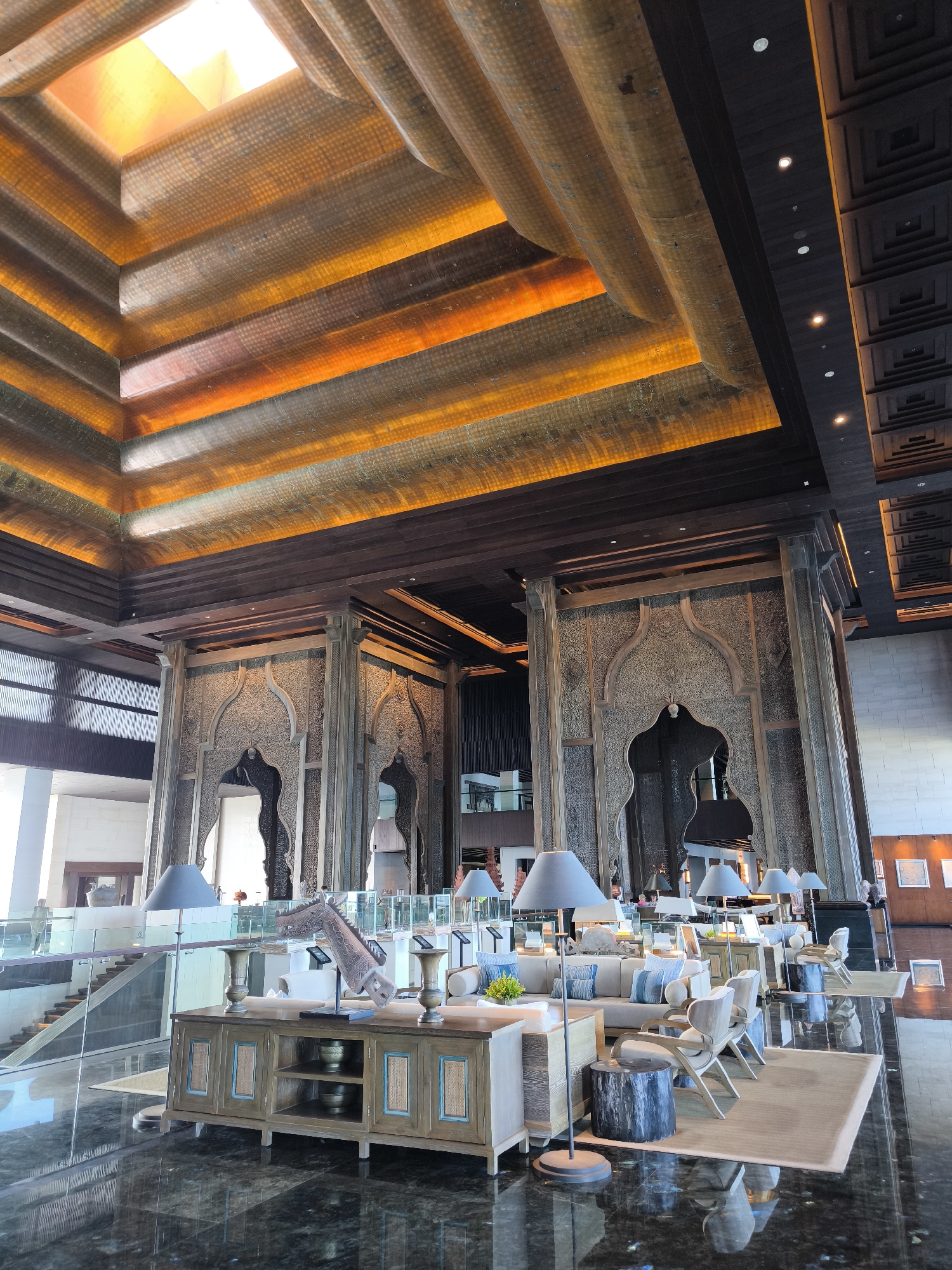 The image depicts the opulent interior of a hotel lobby in Kuta Selatan, Indonesia. The ceiling features a multi-layered design with illuminated amber-colored panels, creating a warm and inviting ambiance. Intricately carved wooden screens with arched openings adorn the walls, showcasing a sophisticated, possibly Middle Eastern-inspired aesthetic.
In the foreground, a polished dark marble floor reflects the surrounding decor. A wooden console table with multiple shelves and cabinets is positioned centrally, displaying decorative vases and a carved wooden horse head sculpture. A grey floor lamp stands beside it.
To the right, a seating area is arranged with plush sofas and armchairs, adorned with light blue and grey cushions. Several matching floor lamps with grey shades are strategically placed around the seating area. A large woven rug anchors the seating arrangement.
In the background, a grand staircase with glass railings ascends to an upper level, further emphasizing the spaciousness of the lobby. Various potted plants and decorative elements are visible throughout the scene, contributing to the overall luxurious atmosphere. Two individuals are partially visible in the background, one standing near the staircase and another seated in the lounge area, suggesting the lobby is in use. The lighting is a mix of natural light from a skylight and artificial lighting from the ceiling and lamps, casting soft shadows and highlighting the architectural details.