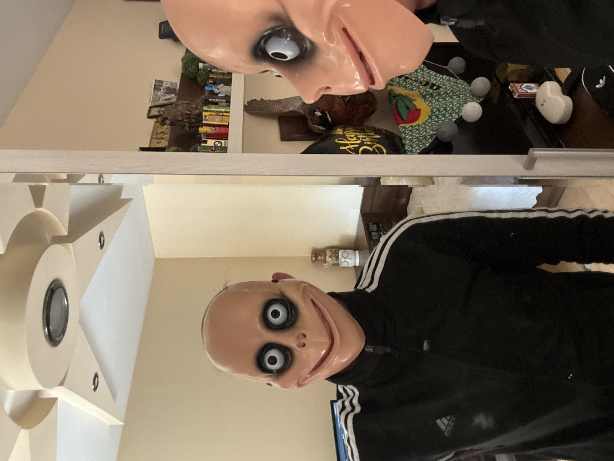 The media file is an indoor photograph, rotated 90 degrees counter-clockwise. It features a person wearing a "Momo" mask and an Adidas tracksuit, captured both directly and as a reflection in a mirror or glass surface.
The central subject is a person wearing a pale, unsettling mask that depicts a bald head with large, bulging black and white eyes and a wide, unnerving smile. The person is dressed in a black tracksuit adorned with distinctive white stripes, consistent with the Adidas brand (the Adidas logo is visible on the lower part of the tracksuit in one of the provided crops).
The scene is an indoor setting, likely a room within a home in Varna, Bulgaria. The background, viewed through the reflection, reveals various household items. These include a mounted, taxidermied bird (possibly a pheasant), stacks of books or small decorative boxes, and a green patterned bag with a fruit design. Near these items, a red and white box (resembling playing cards), three small fuzzy balls, and a white heart-shaped ornament are visible. A black balloon with gold lettering, partially visible as "Happy Birth...", suggests a "Happy Birthday" celebration.
In the non-reflected part of the image, where the person is directly pictured, a light-colored wall serves as the backdrop. A modern, geometric white light fixture with a central circular element is mounted on the wall or ceiling to the left. A small decorative jar or bottle sits on a shelf behind the person's head.
The activity appears to be a casual photo-taking moment, possibly at a party or during a fun event, given the birthday balloon and costume mask. The mask itself conveys a creepy or unsettling emotion due to its exaggerated features.
Visible text includes the "Adidas" logo on the tracksuit and "Happy Birth..." on the balloon.