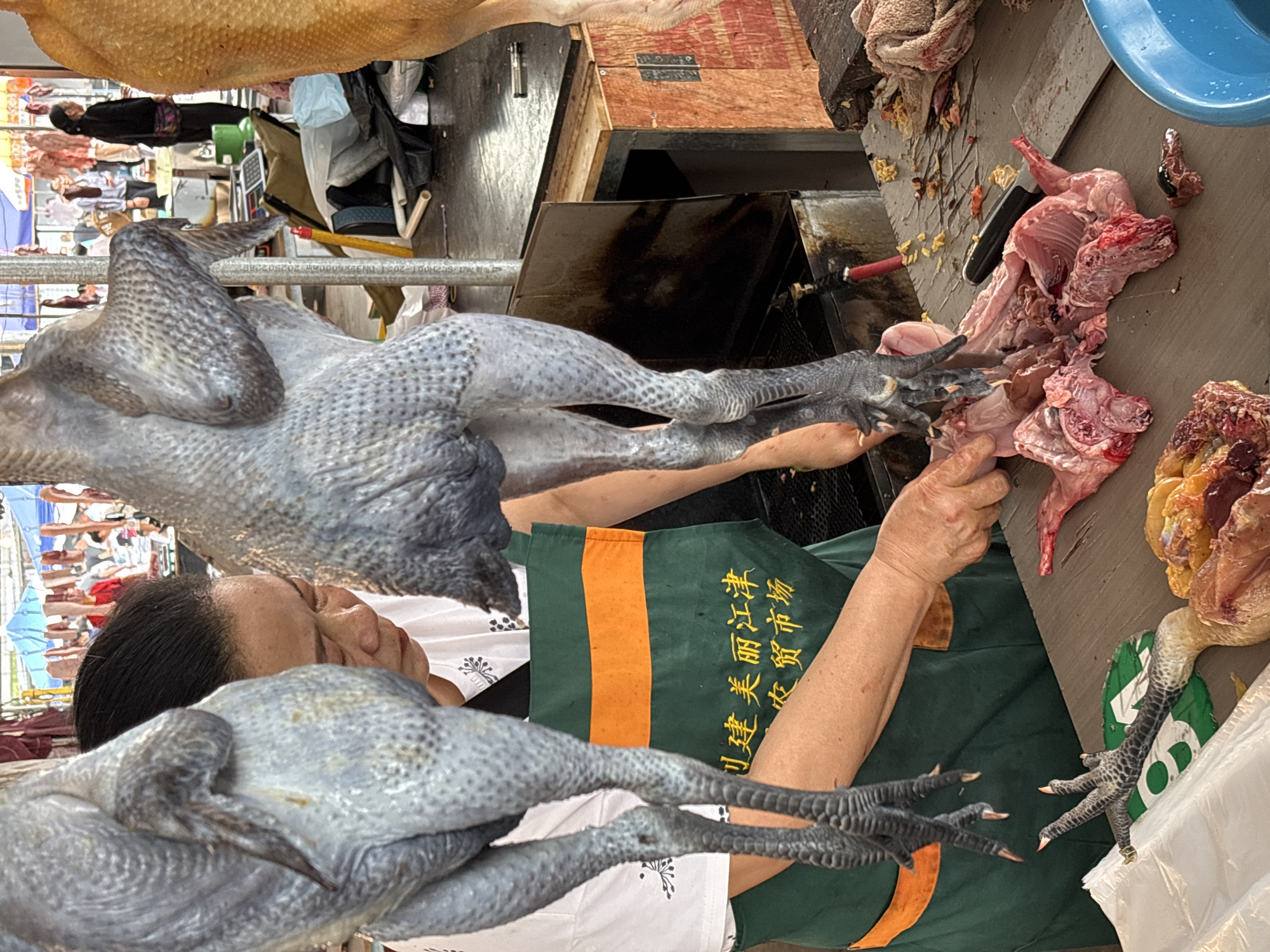 A person wearing a green and orange apron and a white shirt with a black pattern is butchering a black chicken at a market. The butchering surface is gray. A butcher knife lies on the butchering surface. Partially butchered fowl are on the right side of the butchering surface. Two whole black chickens are near the butcher. The background shows a variety of vendors in the market. The text on the apron indicates that the market is located in Jiangjin, China.