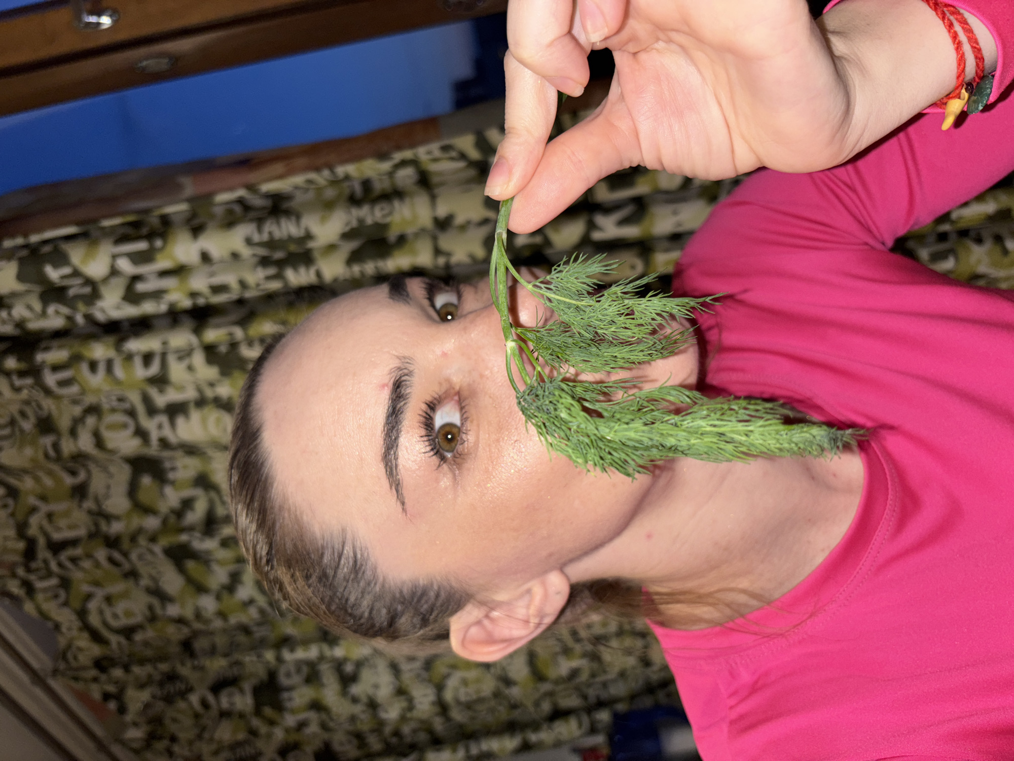 A close-up, eye-level shot shows a woman in Sinyavino, Russia, holding a sprig of dill to her nose. She is wearing a bright pink long-sleeved shirt. Her left eye is wide open and looking directly at the camera, while her right eye is partially obscured by the dill. The dill's feathery fronds are spread across her face, creating a humorous effect. The background is a blur of green and yellow patterned curtains. A window with a dark blue sky is visible to the right of the curtains. The woman has fair skin, brown eyes, and dark blonde hair pulled back in a ponytail. A thin red bracelet with a small yellow charm and green bead is visible on her right wrist.