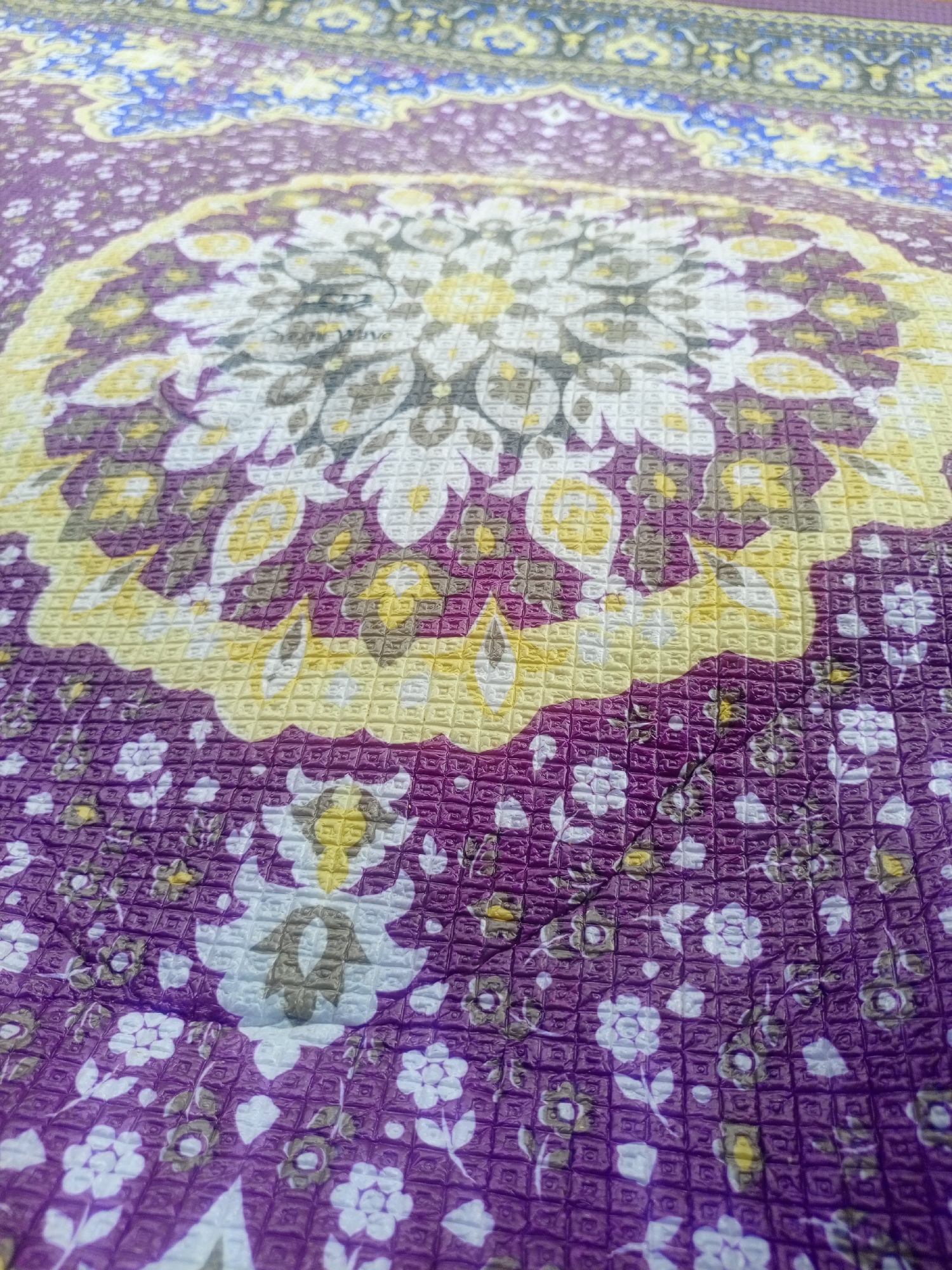 The image shows a close-up, angled view of a textured purple fabric with a floral mandala pattern. The pattern features concentric circles of floral motifs in white, yellow, and grey, set against a background of smaller white and yellow flowers on a purple grid-like texture. There are no people, buildings, or any discernible city elements, making it impossible to determine its location within Jos, Nigeria.