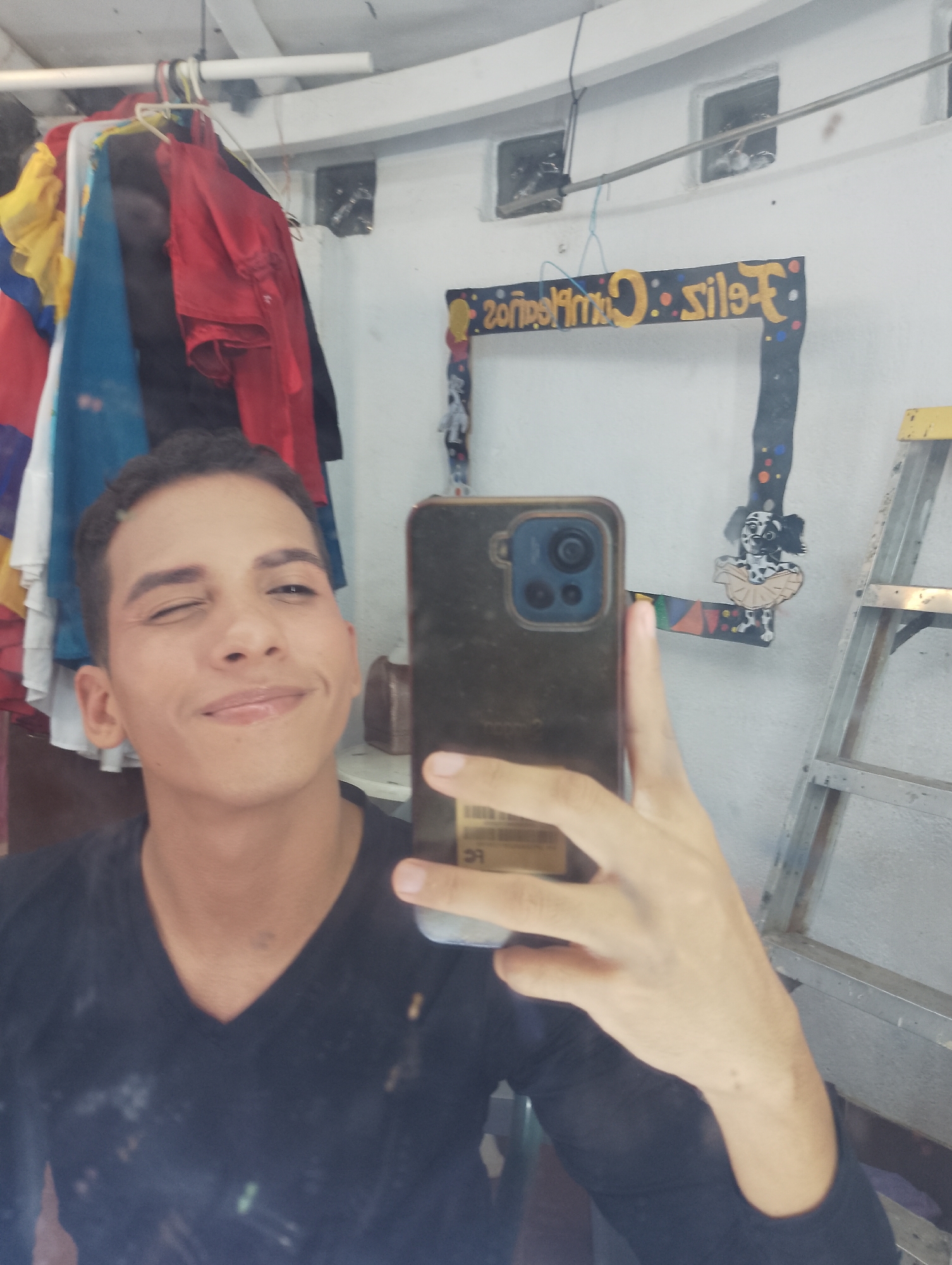 This image, taken in Porlamar, Venezuela, displays a reflection of a young adult male winking at the camera, holding a smartphone to capture the image. The male, positioned centrally, has short dark hair, an olive skin tone, and is wearing a dark V-neck long-sleeve shirt. His left eye is closed in a wink, and a faint smile is present. He holds a dark-cased smartphone with a visible blue camera module in his right hand, covering the lower right portion of his face.
The background, visible through the reflection, reveals an indoor setting. To the left, a white clothes rack holds several garments, including a yellow and blue item, and multiple red shirts. The upper background features a white ceiling with a pipe or beam running across it, and two rectangular wall openings, possibly vents or small windows, are visible above a horizontal metallic bar. To the right of the male, a decorative, dark-colored frame is present, featuring the mirrored text "Feliz Cumpleaños" (Happy Birthday) in yellow letters adorned with colorful dots. A cutout figure of a white dog with black spots, wearing a tutu, is attached to the lower right section of the frame. Further to the right, a silver-colored folding step ladder is leaning against the wall. The reflective surface itself, through which the image is taken, exhibits some smudges and dust.