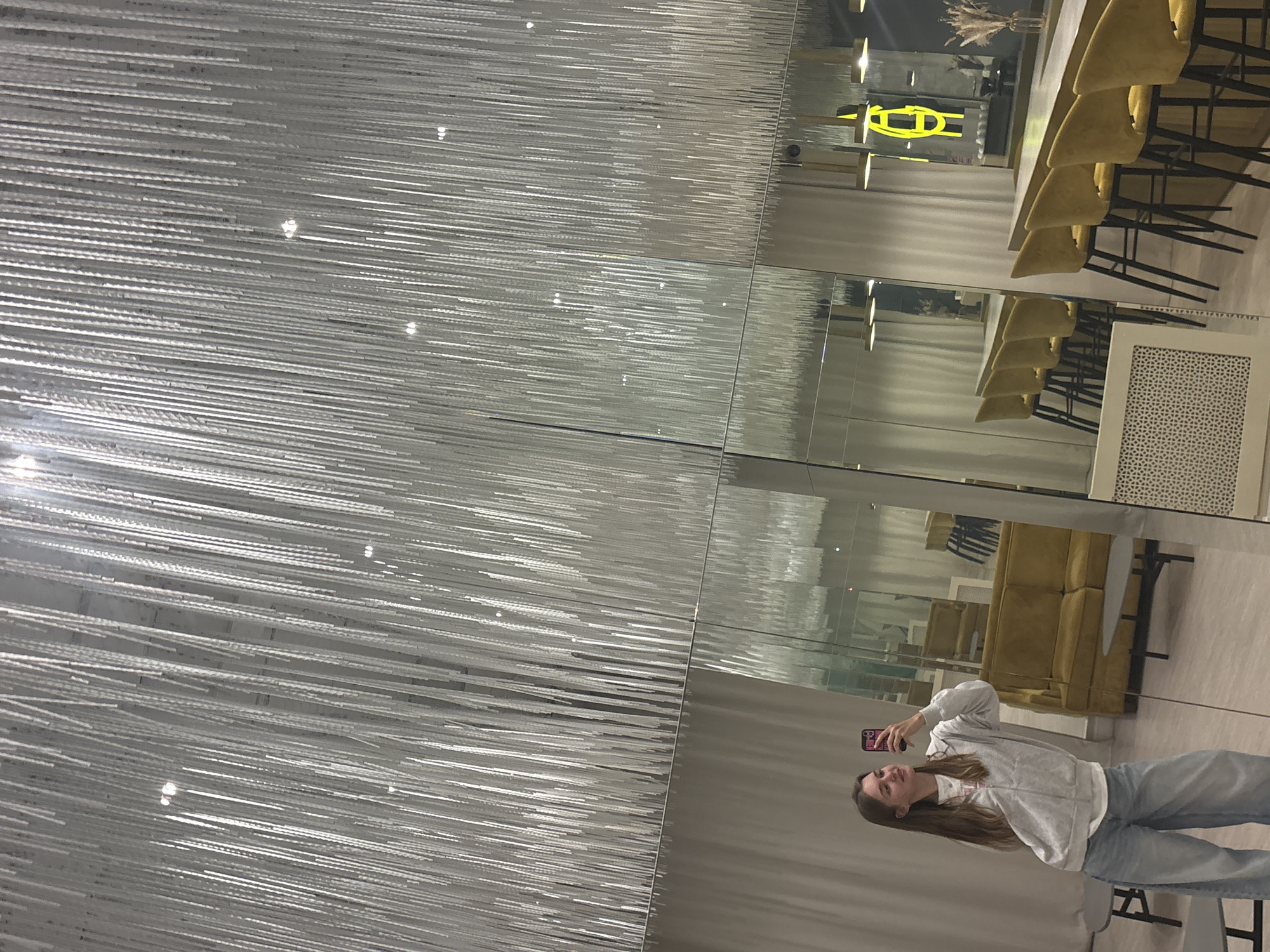 The image depicts a woman taking a picture of herself in a mirrored space. Her reflection is captured in multiple mirrors, showing her holding a phone, wearing a light-colored sweatshirt and jeans. The reflection also shows dining tables and chairs as well as a sofa in the background. The reflection shows the location is in Perm, Russia.