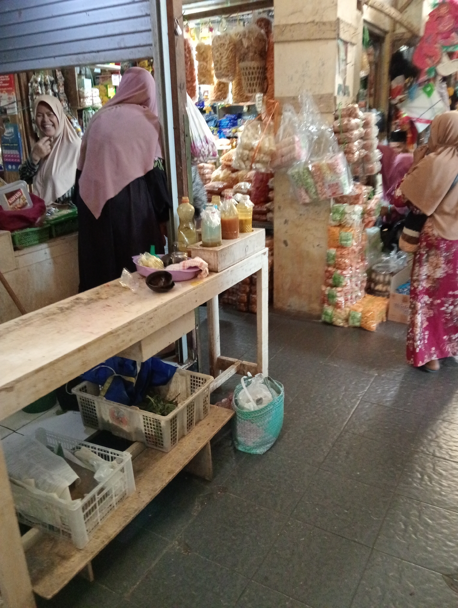 The image depicts a market scene in Grabag, Indonesia. In the foreground, a wooden counter with various items is visible. On the counter, there are two bottles of liquid, a purple bowl containing some food, and a small black bowl. Below the counter, two white plastic crates are placed on a wooden shelf. The left crate contains items wrapped in paper and plastic, while the right crate holds green leafy vegetables. A teal woven basket with a white plastic bag inside sits on the floor to the right of the crates. In the background, two women are present. One woman, on the left, wears a light-colored hijab and is smiling. The other woman, on the right, wears a light brown hijab and a floral patterned dress. Various goods, including packaged snacks and other items, are displayed on shelves and racks behind the women. The market floor is paved with dark tiles.