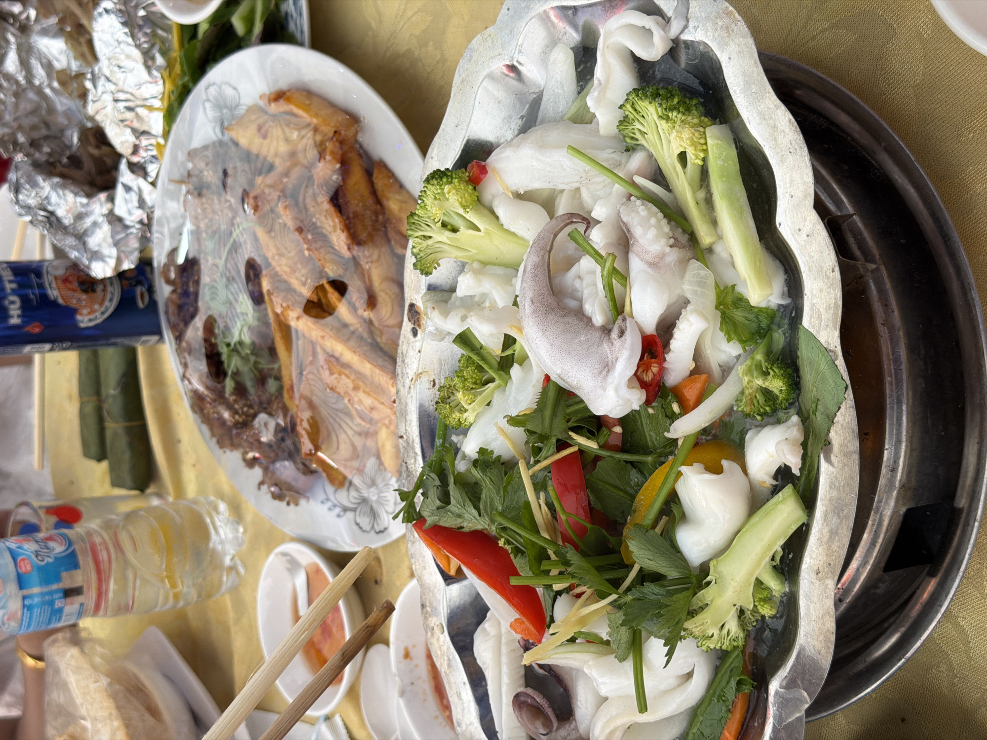 The image captures a vibrant overhead view of a dining table laden with Vietnamese dishes, likely in Tay Son Commune, Vietnam. The central focus is a large, ornate metal platter or pot containing a fresh assortment of ingredients, possibly for a hotpot or steamed dish. This main dish features substantial pieces of white squid, bright green broccoli florets, thin slices of red and yellow bell pepper, slivers of ginger, and various green herbs such as cilantro and parsley. The ingredients appear uncooked or lightly prepared, ready for further cooking at the table.
To the left, a ceramic plate holds several pieces of what appear to be fried or baked goods, possibly fish or snacks, covered with a clear plastic wrap. Adjacent to this, crumpled aluminum foil and some items wrapped in green banana leaves (possibly *banh tet* or a similar snack) are visible.
Beverages on the table include a partially visible blue can with the text "HÔ TÉ" clearly legible, indicating Huda Beer, a popular Vietnamese brand. A plastic water bottle, partially filled, is also present.
The table itself is covered with a golden or light yellow tablecloth. Chopsticks rest on small white dipping bowls, suggesting a communal dining experience. The overall setting is casual, typical of a family meal or a gathering at a local eatery. The bright lighting suggests it is daytime.