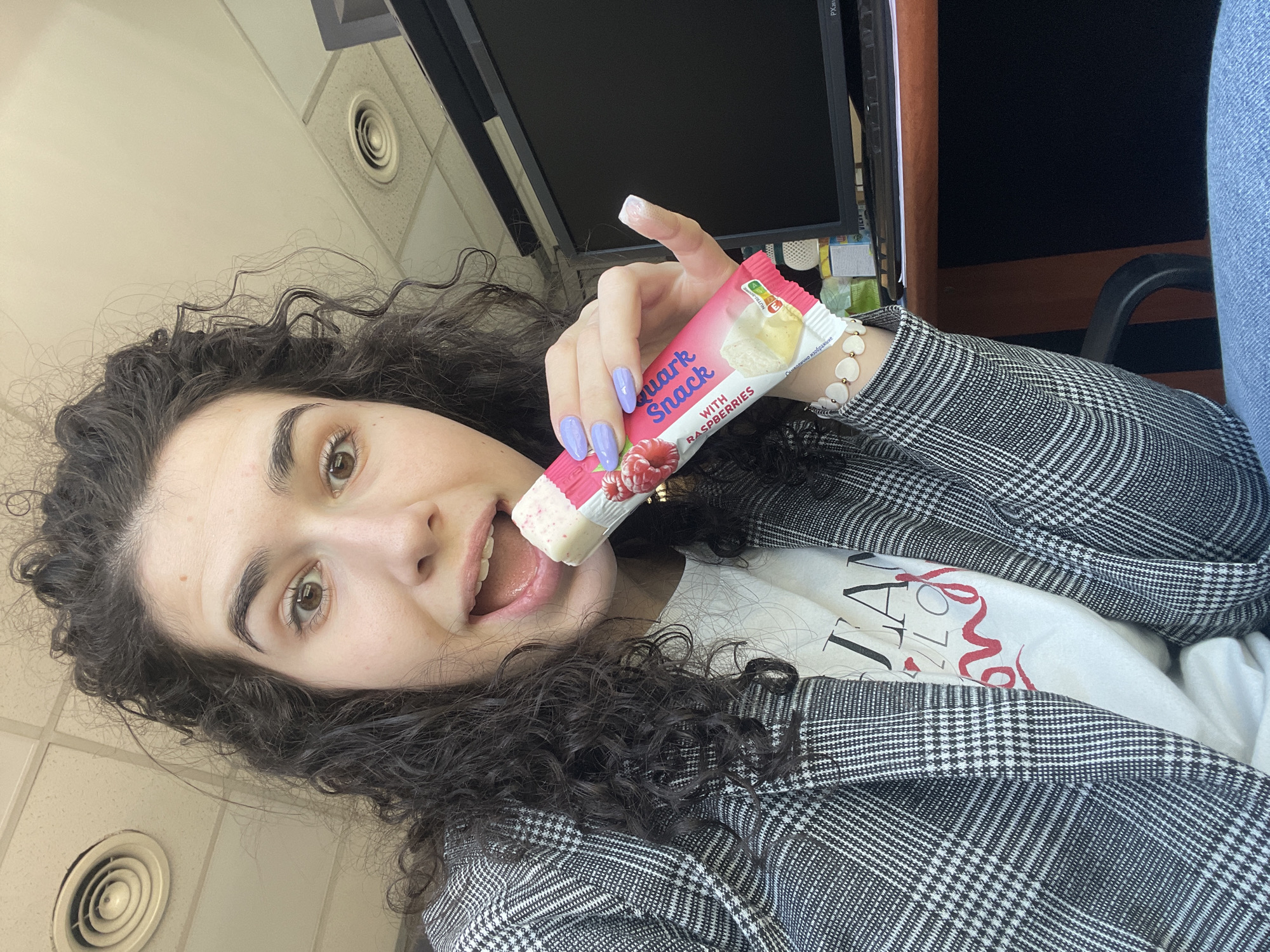 A woman with curly dark hair is holding a partly eaten "Quark Snack with Raspberries" in front of her open mouth. She is wearing a gray plaid blazer over a white t-shirt with red lettering. Her fingernails are painted light purple. She has a bracelet on her left wrist. The background includes a computer monitor, office ceiling tiles with circular vents, a desk, and a chair. There is no indication that the location is in Sofia, Bulgaria.