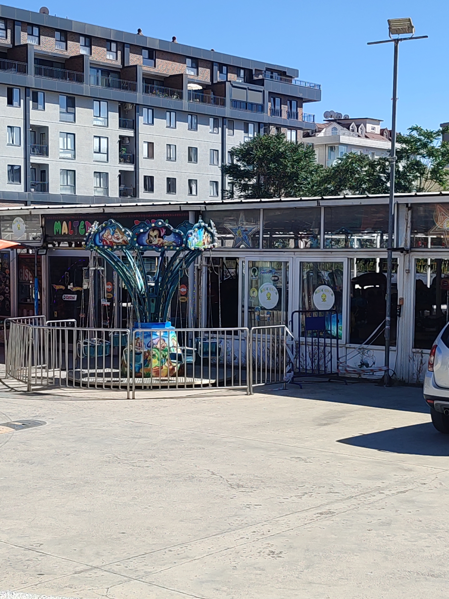 The image shows a children's amusement ride, specifically a swing ride, in Maltepe, Turkey. The ride is blue and green, with chairs hanging from chains. The central pole of the ride is decorated with images of cartoon characters. It is surrounded by a metal fence. Behind the ride, there is a building with large glass windows, suggesting it might be a shop or an arcade. The building's facade is decorated with colorful signage. In the background, a modern apartment building is visible, along with trees and a clear blue sky. A lamppost stands to the right of the amusement ride, and a white car is partially visible in the lower right corner. The ground is paved with concrete, with some tire marks visible. The scene appears to be an outdoor public space.
