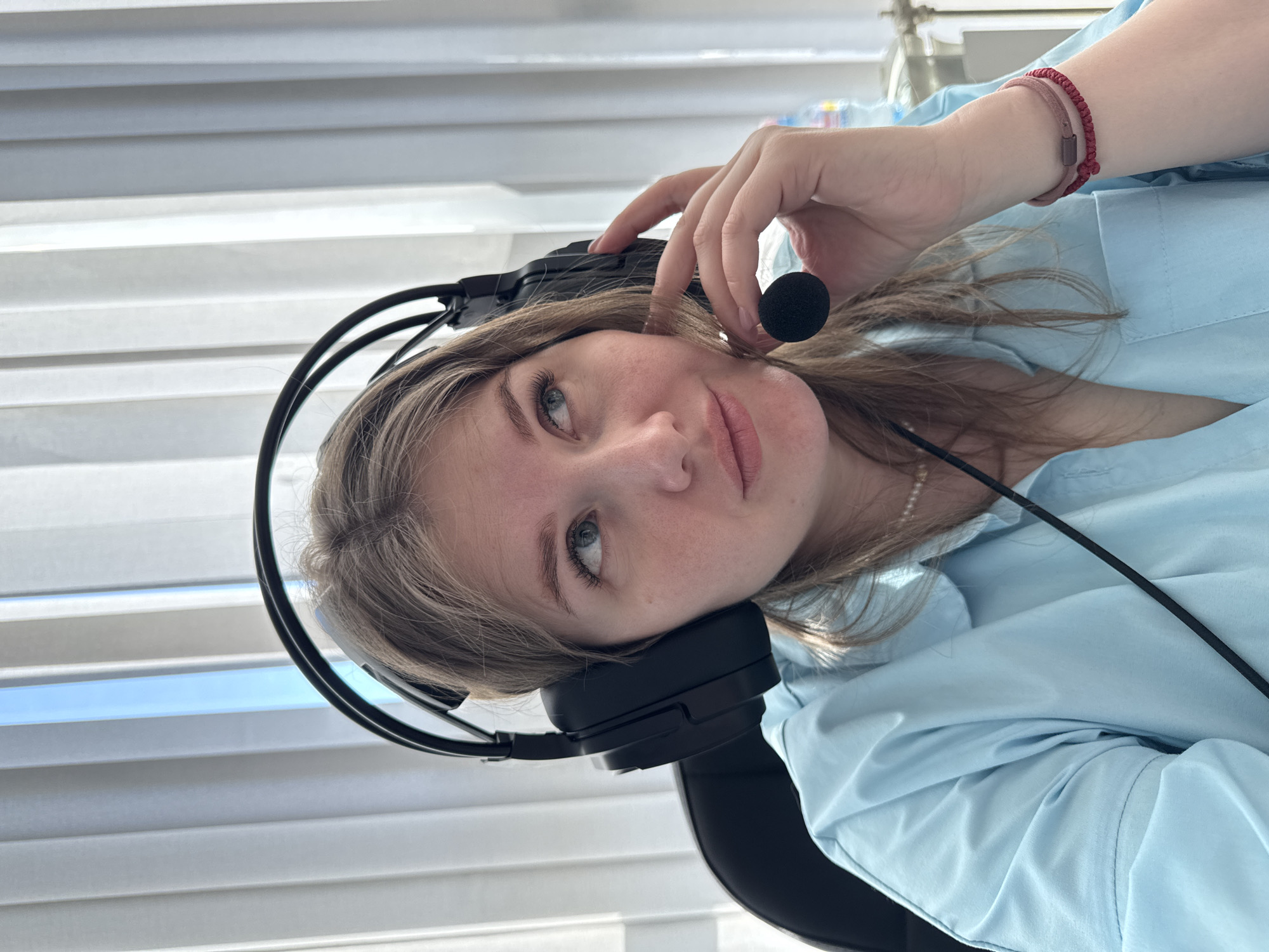 A young woman is depicted in a portrait-style image, rotated 90 degrees counter-clockwise, showing her from the chest up. She has light brown hair and is wearing a light blue long-sleeved collared shirt. A black over-ear headset with a microphone boom is on her head, and her left hand is gently placed near the microphone. Her right wrist features two bracelets: one dark red and braided, the other a lighter red or pink. A subtle pearl necklace is also visible around her neck. She looks directly at the viewer with a calm, slightly contemplative expression.
The background consists primarily of white venetian blinds, suggesting an indoor setting like an office or home office, with natural light filtering in. A portion of a black chair is visible on the right side of the frame. In the upper right corner, slightly out of focus, some water bottles are faintly discernible. The woman's attire and the headset imply she is engaged in an activity such as remote work, online communication, gaming, or studying. The lighting suggests it is daytime. There is no visible text in the image.