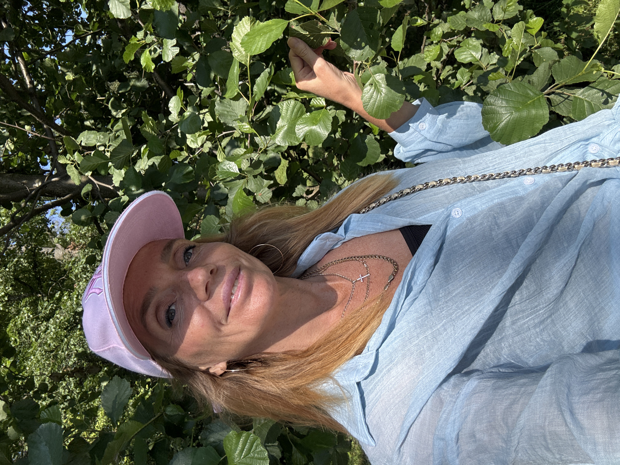 A woman with blonde hair, wearing a light blue collared shirt, a black tank top, and a pink baseball cap, is captured in a close-up, slightly tilted shot. She is outdoors, surrounded by lush green foliage and tree branches, suggesting a natural setting like a park or garden. Sunlight dapples through the leaves, casting shadows and highlights on her face and clothing, indicating it is daytime with bright sunshine. The woman is looking towards the camera with a gentle smile, her eyes are blue and her lips are glossy pink. She is wearing a necklace with a cross pendant and hoop earrings. A bag with a chain strap is slung across her shoulder. The overall mood is relaxed and pleasant, possibly a candid moment captured on a sunny day.