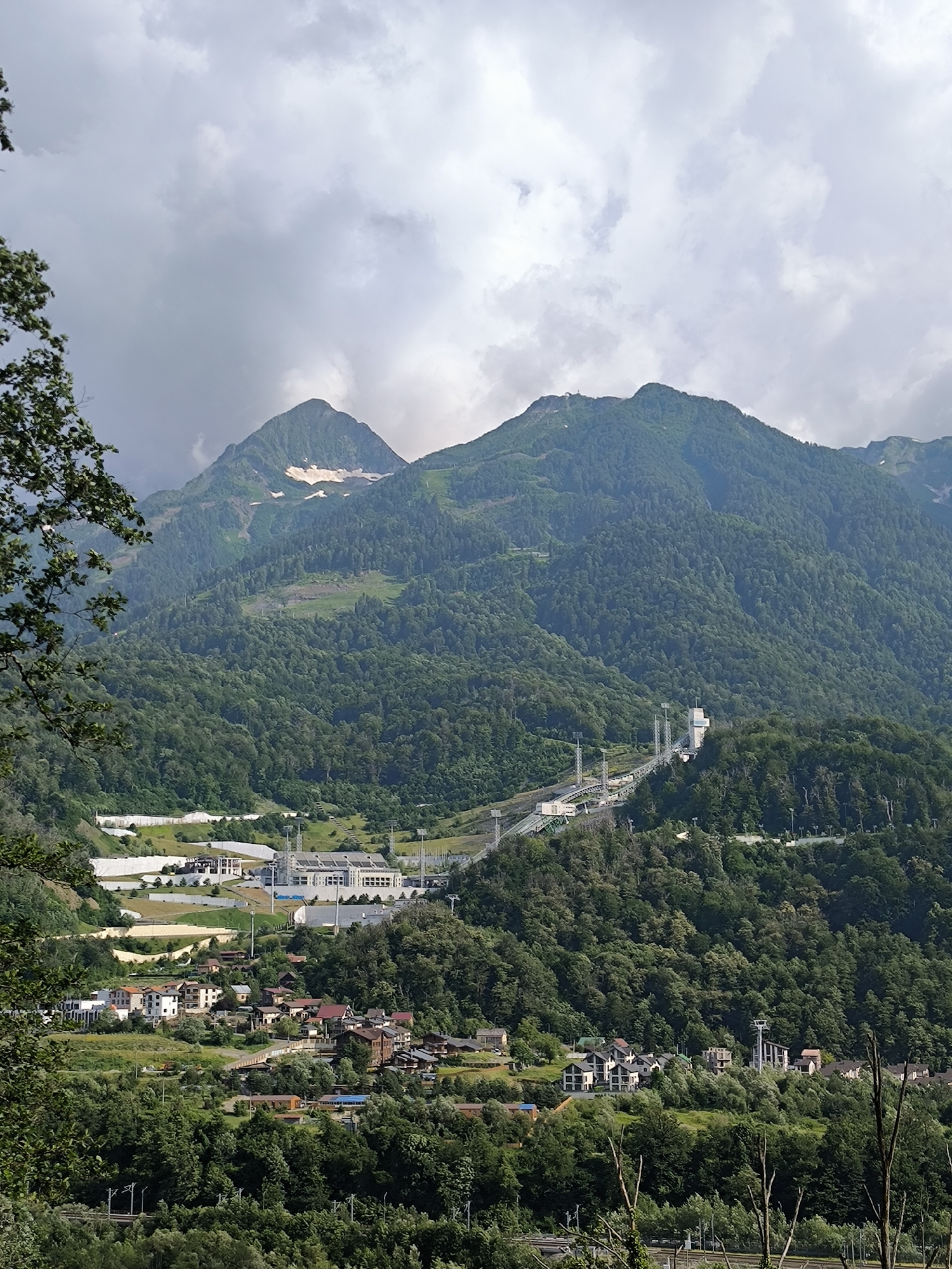 The image depicts a mountain landscape in Krasnaya Polyana, Russia, under a cloudy sky. In the foreground, dense green trees frame the scene. The midground features a town with houses nestled on a hillside, surrounded by greenery. Further up the slope, several modern buildings, possibly sports facilities or infrastructure, are visible, including a large stadium with tiered seating. A ski lift or cable car system extends up the mountain, indicated by poles and cables. The background is dominated by two large, tree-covered mountains, with patches of snow visible on their upper slopes. The overall impression is a developed mountain resort area in a natural setting.