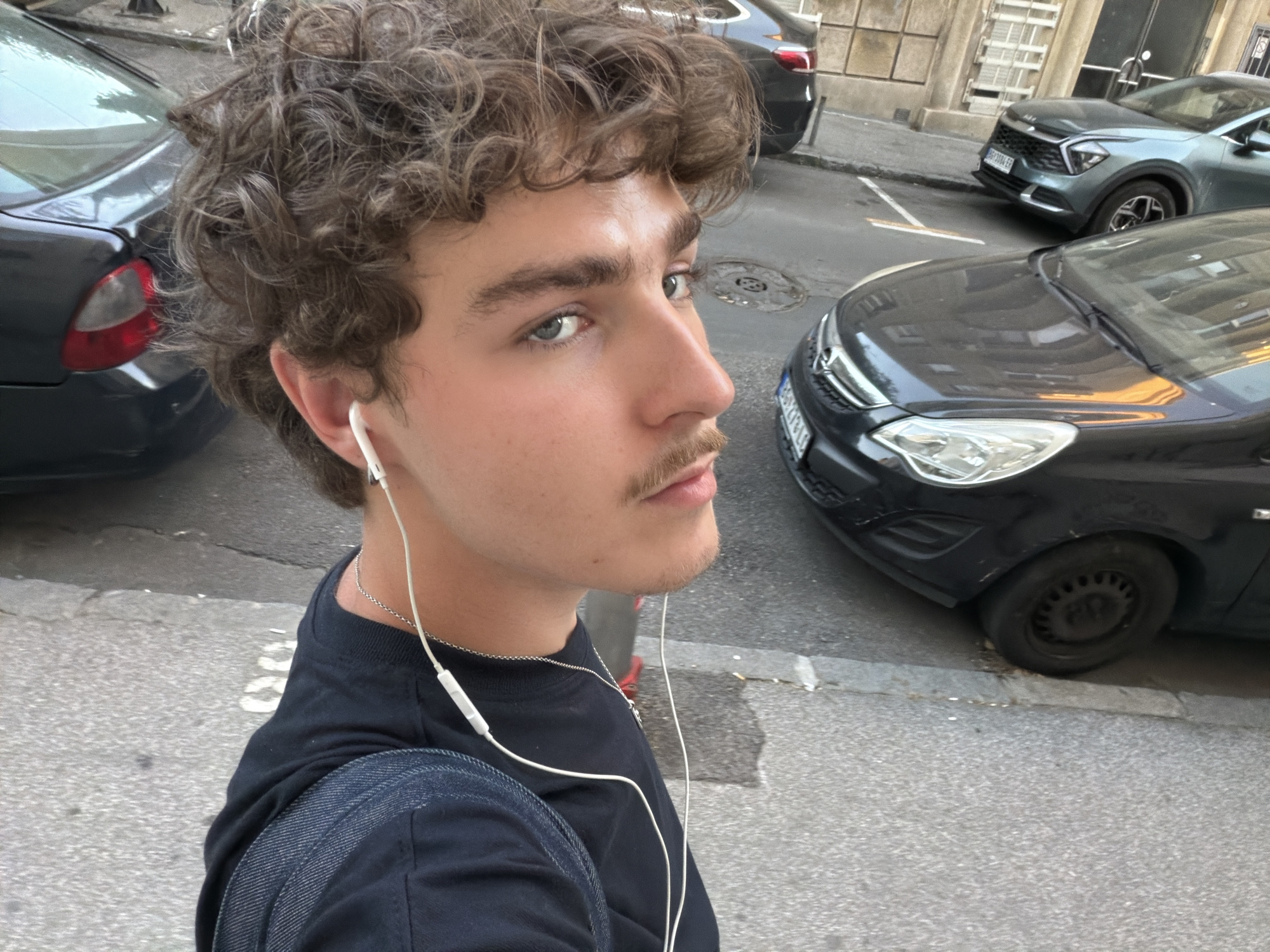 A young man with curly brown hair, blue eyes, and a trimmed mustache stands on a street in Vracar Urban Municipality, Serbia. He wears a black t-shirt, a denim backpack over one shoulder, and white wired earbuds. He is looking towards the viewer. Cars are parked alongside the curb behind him. The street surface is paved with asphalt. A building facade can be seen in the background.