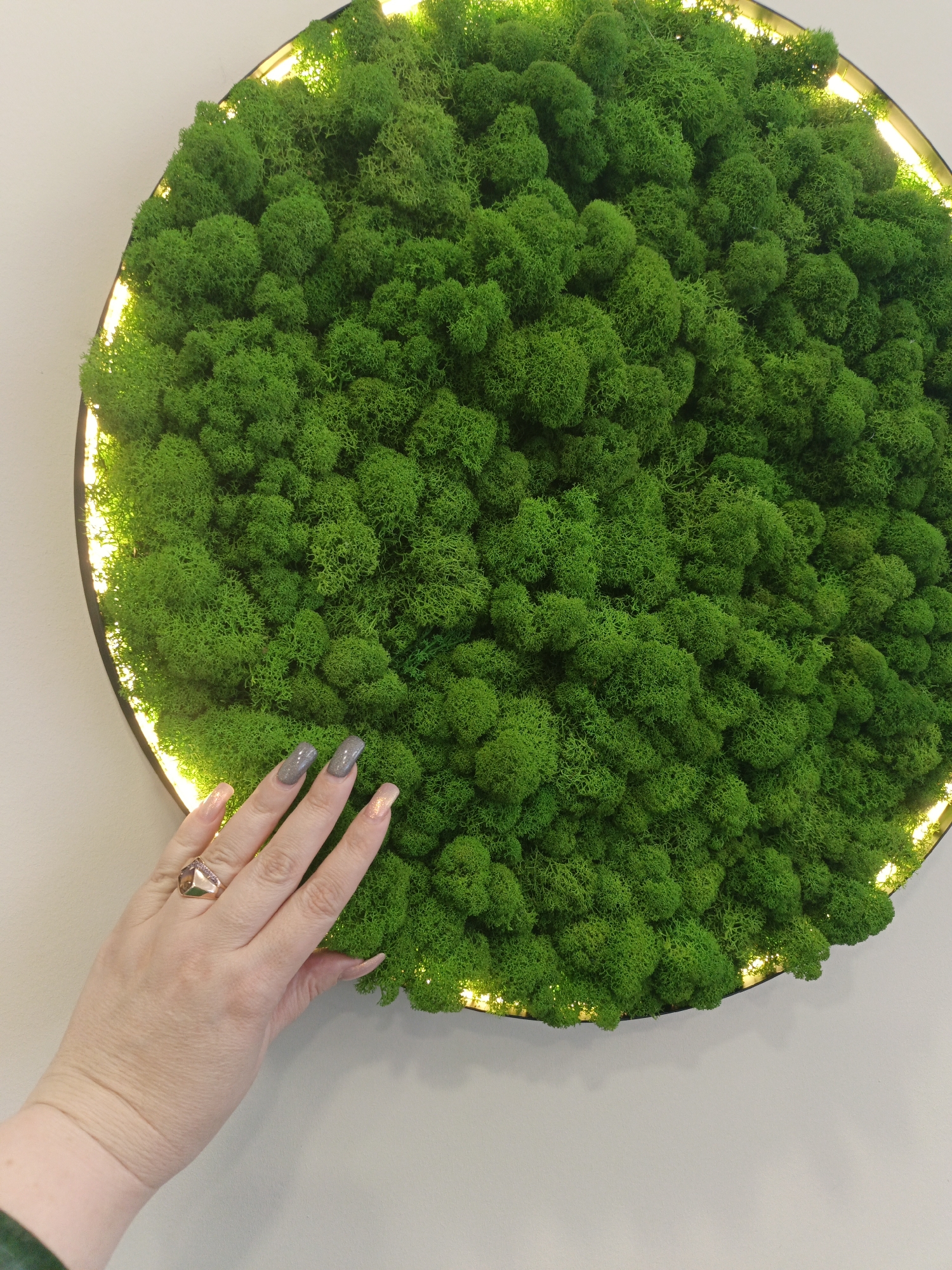 A close-up shot shows a hand resting on a circular panel of bright green moss. The panel has an illuminated edge that casts a warm light around the moss. The hand features long fingernails, some painted gray and some with a pale pink hue. A ring adorns the ring finger. The background is a plain, light-colored wall. The photo's location, Irkutsk, Russia, is only provided as context, not visible elements in the image.